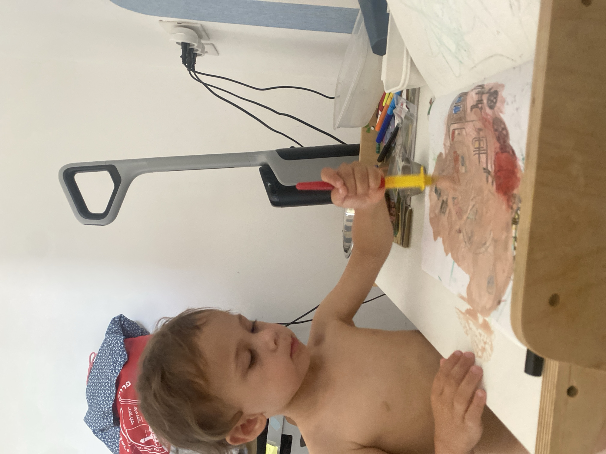 A young, light-skinned child, appearing to be male, is seated at a light-colored table, shirtless and with short, light brown hair. The child is intently focused on an art activity, holding a red and yellow painting tool in their right hand, applying a light brown, paste-like substance, likely paint, onto a white piece of paper. The paper exhibits areas of the brown substance, some red color, and faint blue and black markings beneath the primary layer. The child's left hand rests gently on the paper's surface.
Clustered on the table near the child are various art supplies, including several colored markers, a small metallic bowl, and two white rectangular plastic containers. A light-colored wooden structure, possibly part of a child's art desk or easel, frames the bottom of the composition.
The background consists of a plain white wall. Standing upright against the wall is a grey and black cleaning appliance, identifiable by its handle and main body. To its right, a white electrical wall outlet is visible, from which multiple black electrical cables extend via a multi-plug adapter. In the upper left background, a red bag with partially visible white Hebrew text, "תמיד לטייל על פי דרכך" (always travel your way), is stacked beneath a blue and white patterned fabric item. A vertical strip of light blue fabric is visible on the far right.
This interior scene takes place within a building located in Kiryat Bialik, Israel.