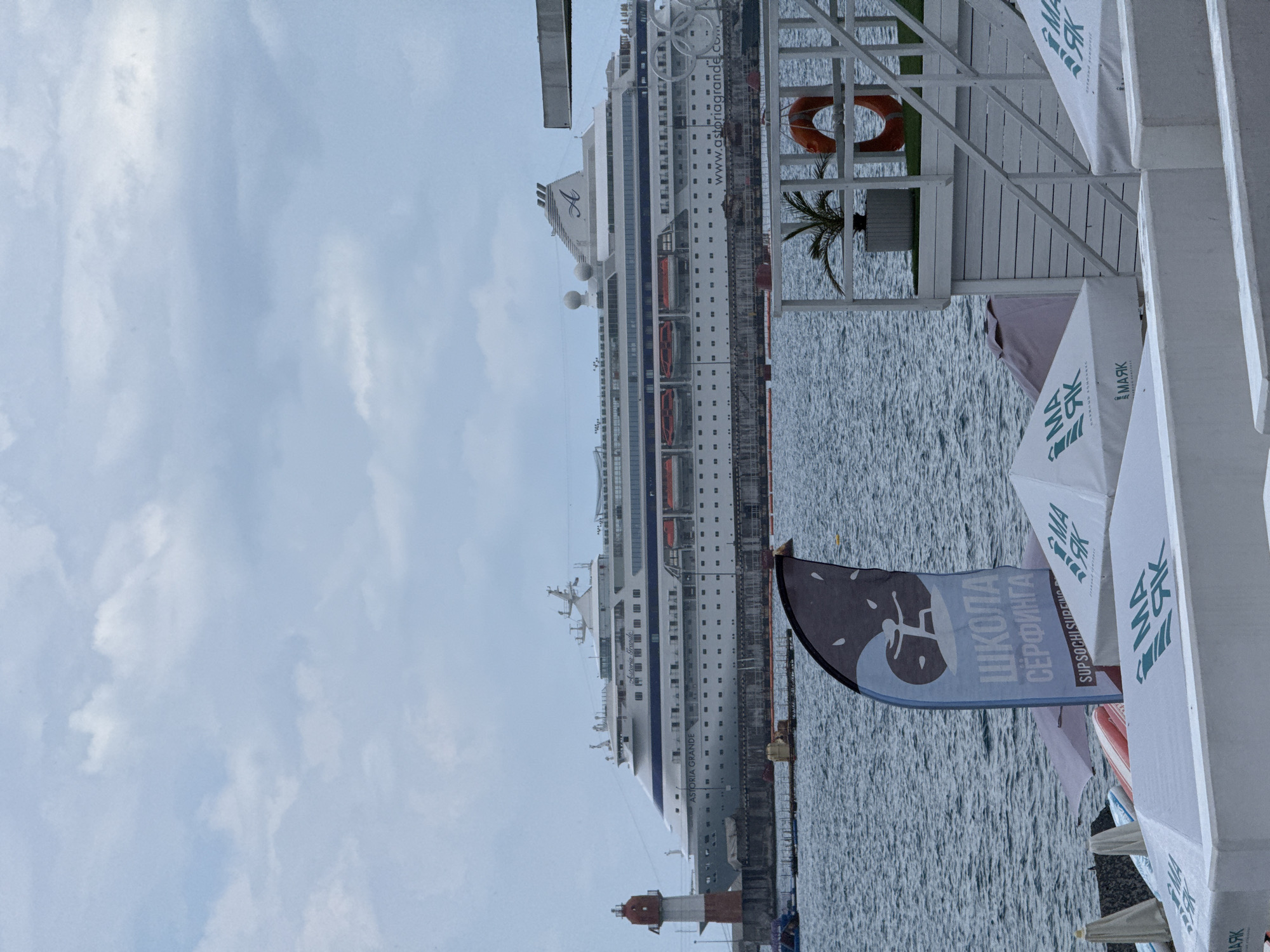 A large white cruise ship, the "Astoria Grande," is docked at the port of Sochi, Russia. The ship has multiple decks and a blue stripe running along its hull. To the left of the ship is a red and white lighthouse. In the foreground, a blue and black flag with a surfing graphic and Russian text that reads "ШКОЛА СЁРФИНГА" (Surf School) is visible. Several white umbrellas with green lettering are also present. The scene is set against a cloudy sky and the water of the Black Sea.