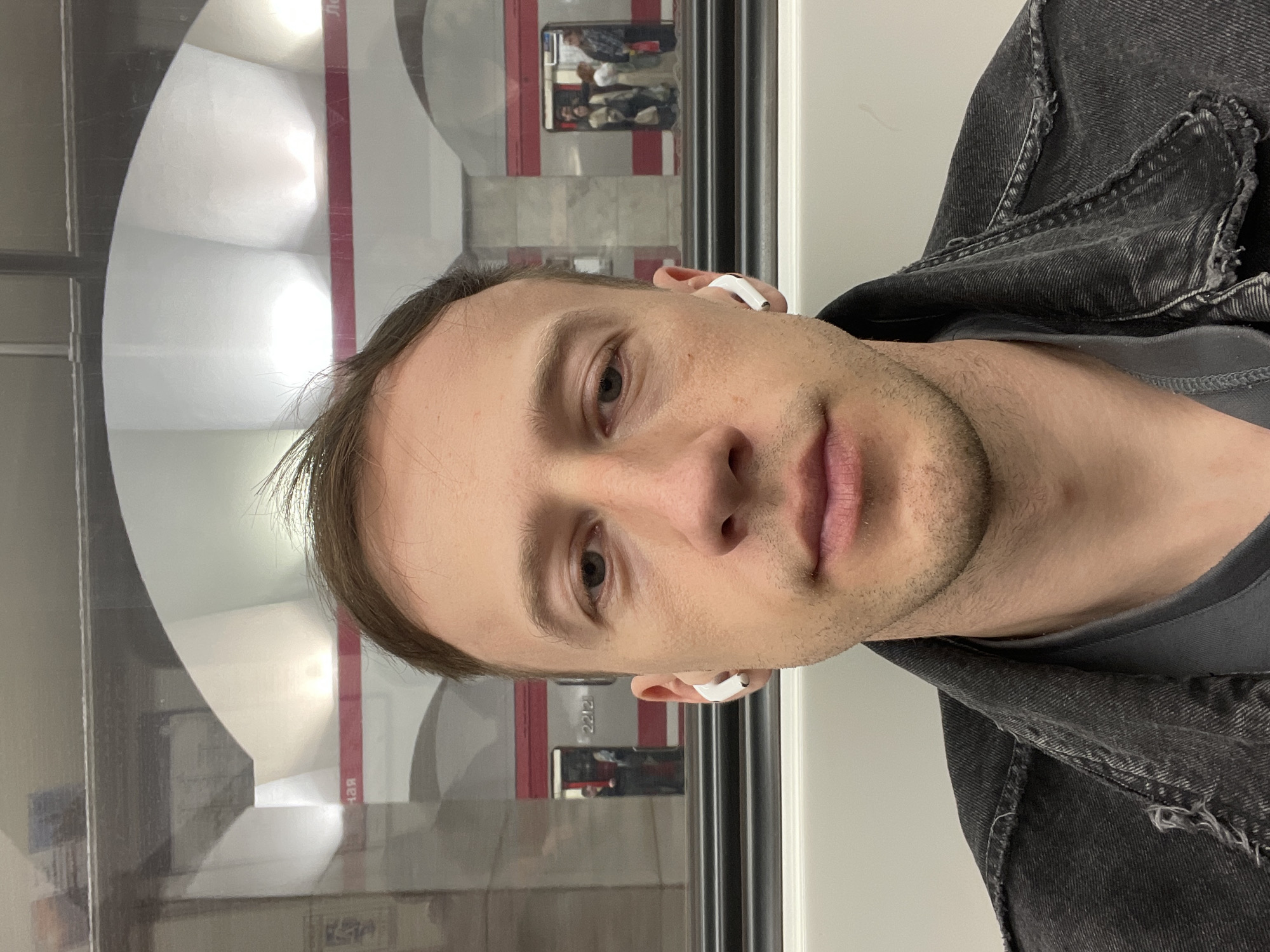 A Caucasian male with short brown hair and light skin is visible in the image, wearing white earbuds in both ears. He is wearing a dark gray t-shirt and a black denim jacket with frayed edges. The background features the interior of a public transportation vehicle, specifically a subway car, with a window showing the platform. The platform has distinctive round, vaulted architectural elements with red stripes, indicating it is a station in the Saint Petersburg Metro system. The image is taken at an angle, suggesting the subject is holding the camera or phone and is situated either sitting or standing. Reflections in the window show activity on the subway platform, implying people are present.