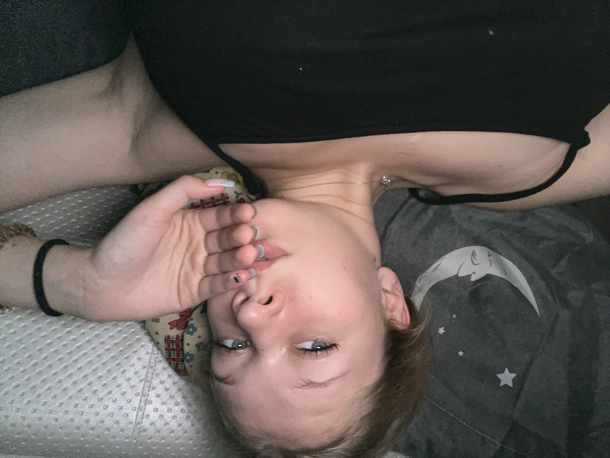 A young Caucasian female is lying down, facing upwards and towards the camera. Her head is resting on a pillow with a colorful floral pattern. She has blonde hair, blue eyes, and her right hand is covering her mouth. She is wearing a black tank top and a thin silver necklace with a pendant. Her left arm is bent, with her hand resting on her chest, and a black bracelet is visible on her right wrist. The background includes a gray blanket with a crescent moon and stars pattern. The scene appears to be indoors, possibly in a bedroom. There are no discernable landmarks or objects that would indicate a specific location in the city of Perm, Russia.