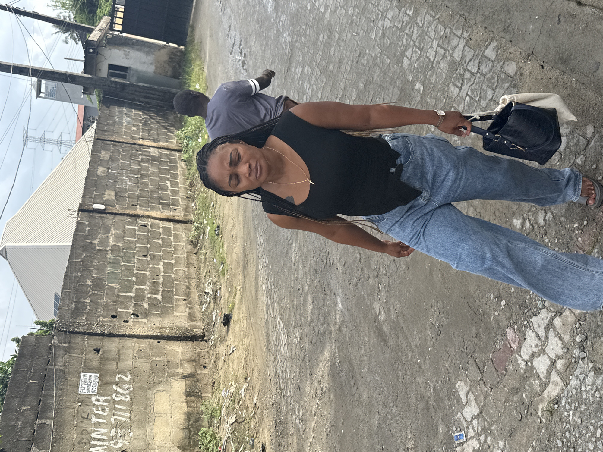 The image shows a woman standing on a cracked, concrete or paved surface alongside a weathered, brick wall in an urban area, likely in Ado, Nigeria. The woman is wearing a black tank top, light blue jeans, and sandals. She is holding a black purse with a white cloth bag protruding from it. A man in a gray shirt is visible in the background. Graffiti is present on the brick wall, with faded writings and a small advertisement sticker mentioning cleaning services and a phone number. The background includes a simple, light-colored building and power lines. The lighting suggests it is daytime.