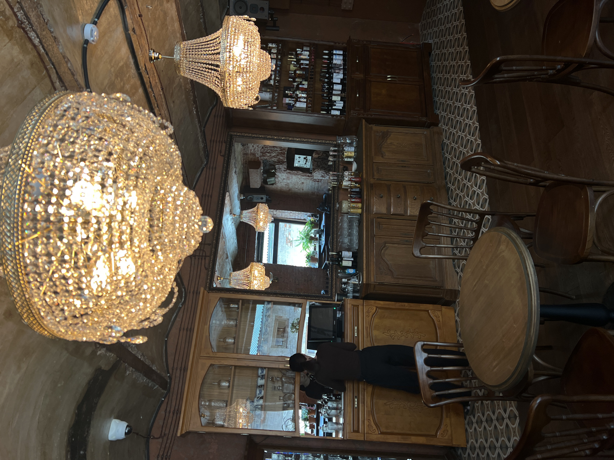 The image shows the interior of a bar, likely in Vladivostok, Russia. Two large crystal chandeliers hang from the ceiling. A back bar is present, with cabinets stocked with bottles of alcohol and glassware. A woman stands at the bar, possibly a worker. A round table and wooden chairs sit in the foreground. The floor is tiled with a patterned design. A mirror behind the bar reflects another part of the room.