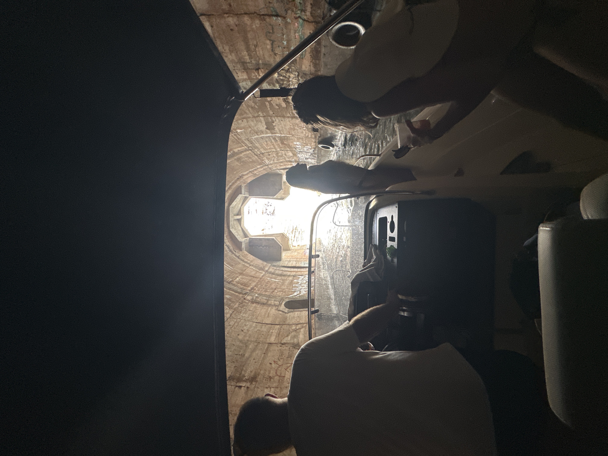 The image captures a boat navigating through a stone tunnel, likely part of the Kumbor fortress in Montenegro. The tunnel's arched walls are visible, showing signs of age and graffiti. Sunlight streams in from the far end, creating a dramatic contrast with the shadowed interior of the boat. Three individuals are present: a man at the helm, facing away from the camera and steering the boat; a woman silhouetted against the light at the front of the boat, facing forward; and another individual, partially visible from the back and side, sitting on the right side of the boat. The water appears calm, reflecting the tunnel's architecture.