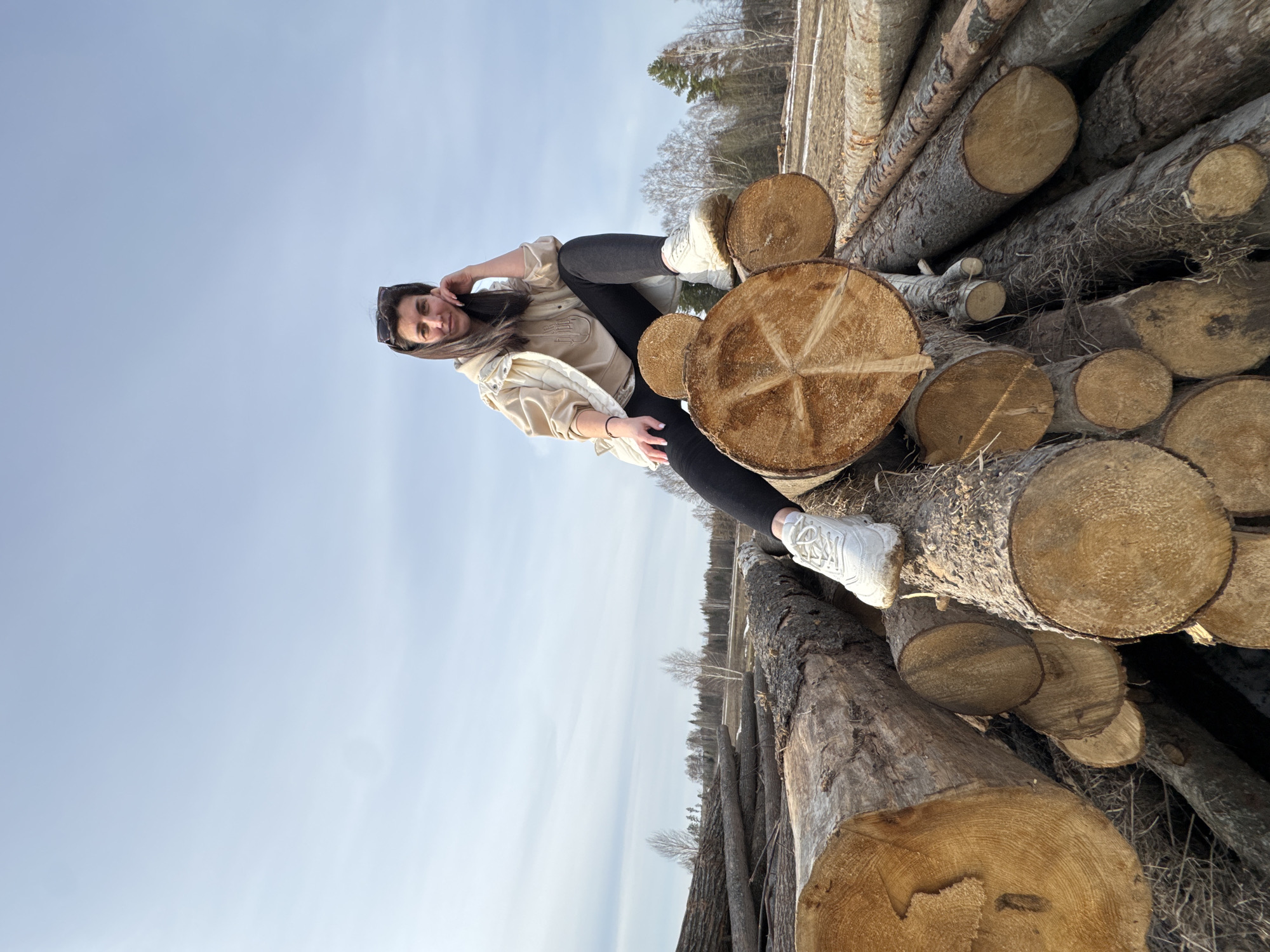 A woman with long brown hair and sunglasses is reclining on a stack of logs. She is wearing black leggings, a light beige jacket, and white sneakers. The logs are various sizes and appear to be freshly cut. The background consists of a clear blue sky and a distant line of trees. The location is Van'ki, Russia.