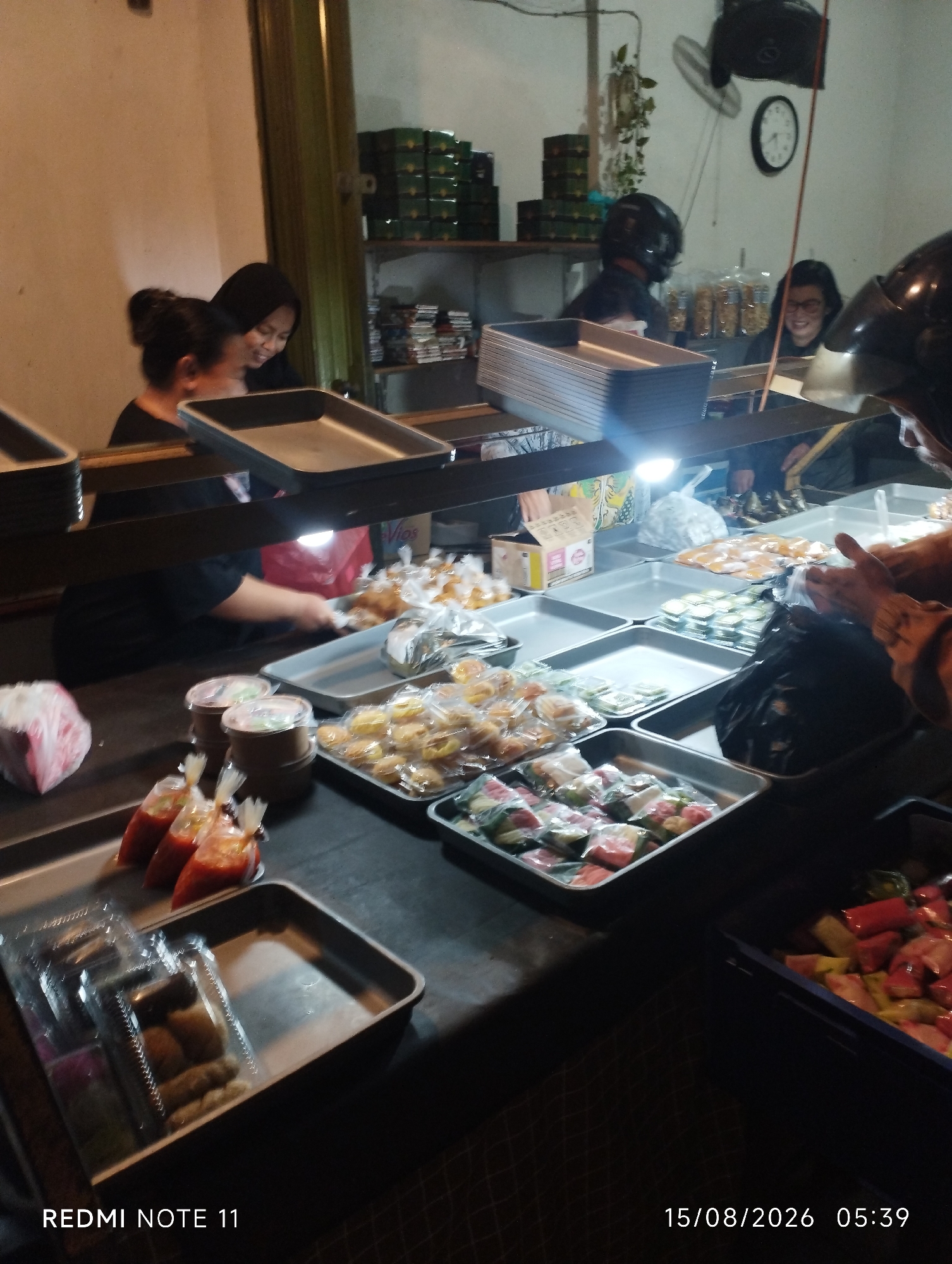 The image depicts an indoor scene at what appears to be a food stall or small eatery, possibly in Karawang, Indonesia, given the timestamp. In the foreground, several trays are visible, containing various food items. One tray holds small, individually packaged snacks, some appearing to be fried or crispy. Another tray contains items wrapped in plastic, possibly some form of dessert or sweet. There are also three plastic sachets filled with a red sauce, likely a condiment.
Behind these food displays, several individuals are present. Two women are seen behind a counter, one with her back to the camera and the other facing sideways, both appearing to be interacting with the food or customers. Further back, a person wearing a helmet and a dark jacket is partially visible, suggesting they might be a customer or worker on a motorcycle. Another individual wearing glasses is also visible in the background, smiling.
The environment suggests a bustling, potentially nighttime operation, with overhead lighting illuminating the food and counter area. Shelves in the background are stocked with boxes. The overall impression is of a casual, street-food setting.