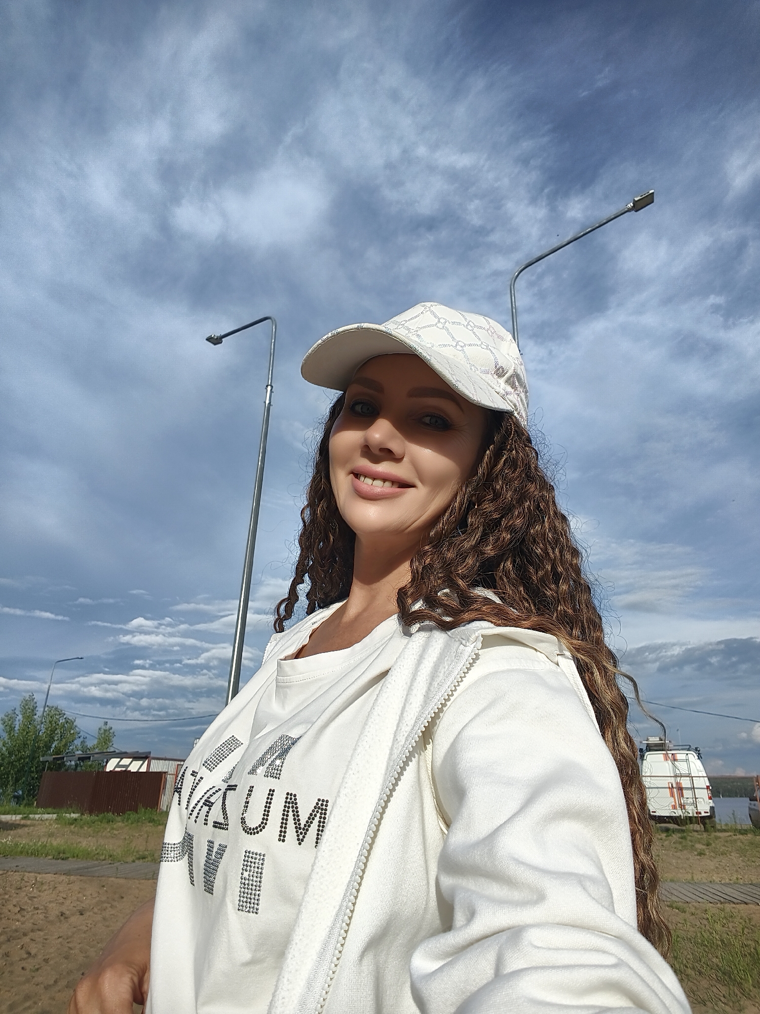 A woman with long, curly brown hair, wearing a white baseball cap and a white zip-up hoodie over a white t-shirt, poses for a selfie outdoors under a cloudy sky. The t-shirt has "AMUSEUM" printed on it in what appears to be rhinestones or reflective material. In the background, two streetlights are visible, along with a wooden fence, some trees, a small van, and a body of water. The location is identified as Chaikovsky, Russia. The scene suggests an outdoor, possibly urban or suburban, setting on a day with mixed cloud cover.