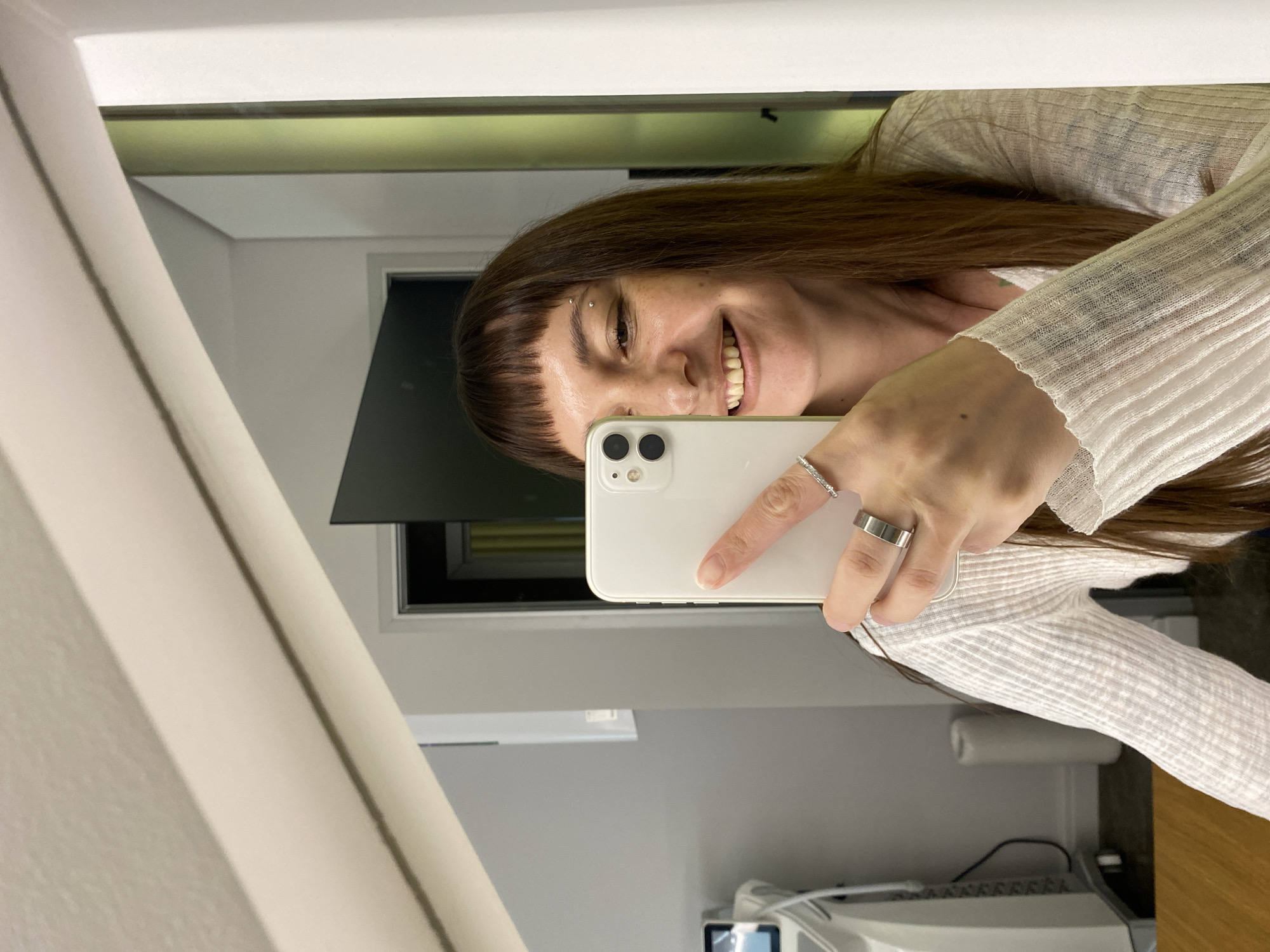 An indoor shot shows a woman taking a mirror selfie. The woman is facing the mirror, holding a white smartphone in her right hand, with her index finger on the screen. She is smiling, with her teeth visible. She has short bangs and long brown hair. She is wearing a white, ribbed long-sleeved shirt. She has two rings on her right hand and an eyebrow piercing. The background of the mirror shows a room with light gray walls, a doorway with a dark green door, and a white rolling object. To the left of the frame, a piece of medical or aesthetic equipment with a screen is visible. The scene is indoors, likely in a private residence or a business establishment. There is no indication of this location being in Votkinsk, Russia.