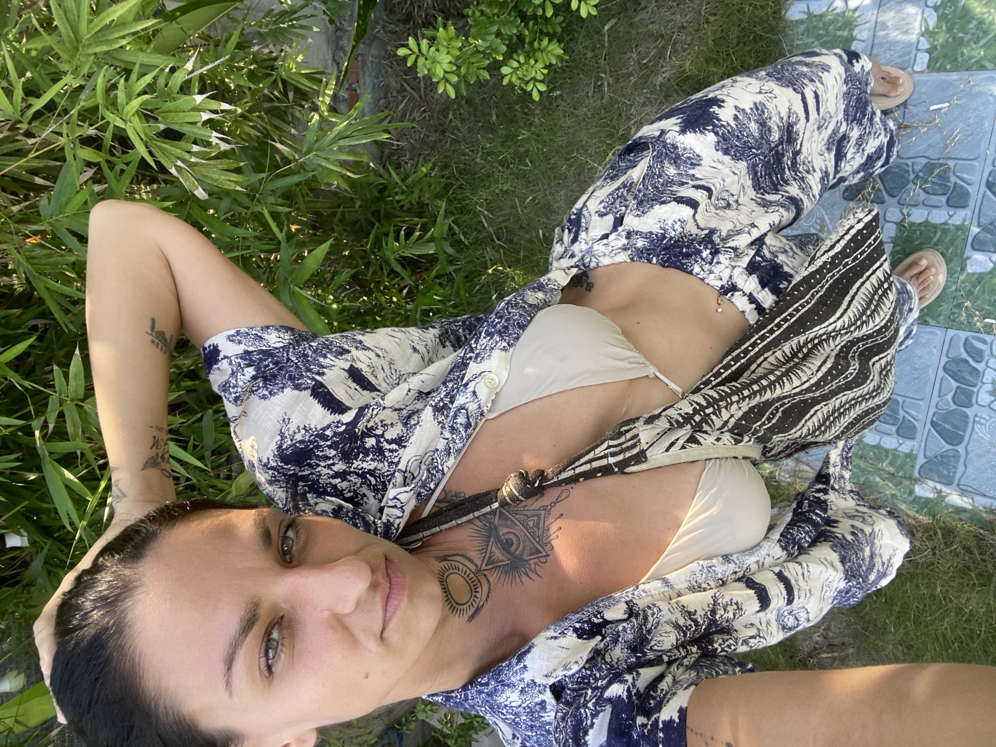 A woman with dark hair and green eyes is taking a selfie outdoors, with lush green foliage in the background. She is wearing a patterned blue and white button-down shirt open over a beige bikini top, and matching patterned pants. A dark and white patterned bag is slung across her chest. Tattoos are visible on her chest, neck, and left arm. She is standing on a pathway made of tiled squares, with grass and more foliage visible around her. The scene appears to be a sunny day in Mũi Né, Vietnam.