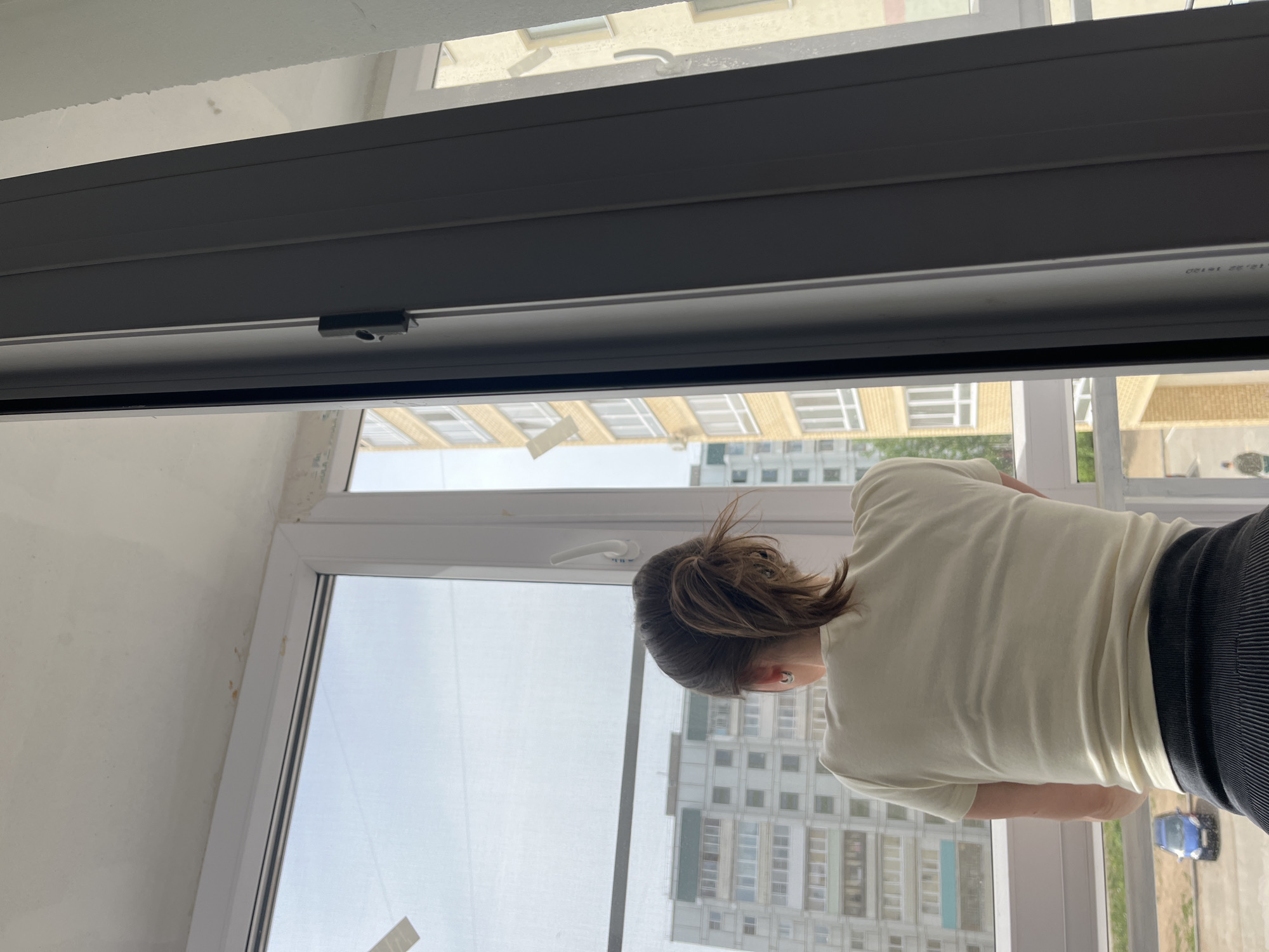 The image depicts a person looking out of a window in a building, possibly an apartment complex, in Chaikovsky, Russia. The individual has brown hair pulled back and is wearing a light-colored t-shirt and dark leggings. The window offers a view of other apartment buildings, indicating an urban residential area. A parked blue car is seen in the distance. The window frame is a light color, and a portion of the window hardware is visible. The building in which the person is located is likely undergoing renovation, based on the visible unfinished wall.