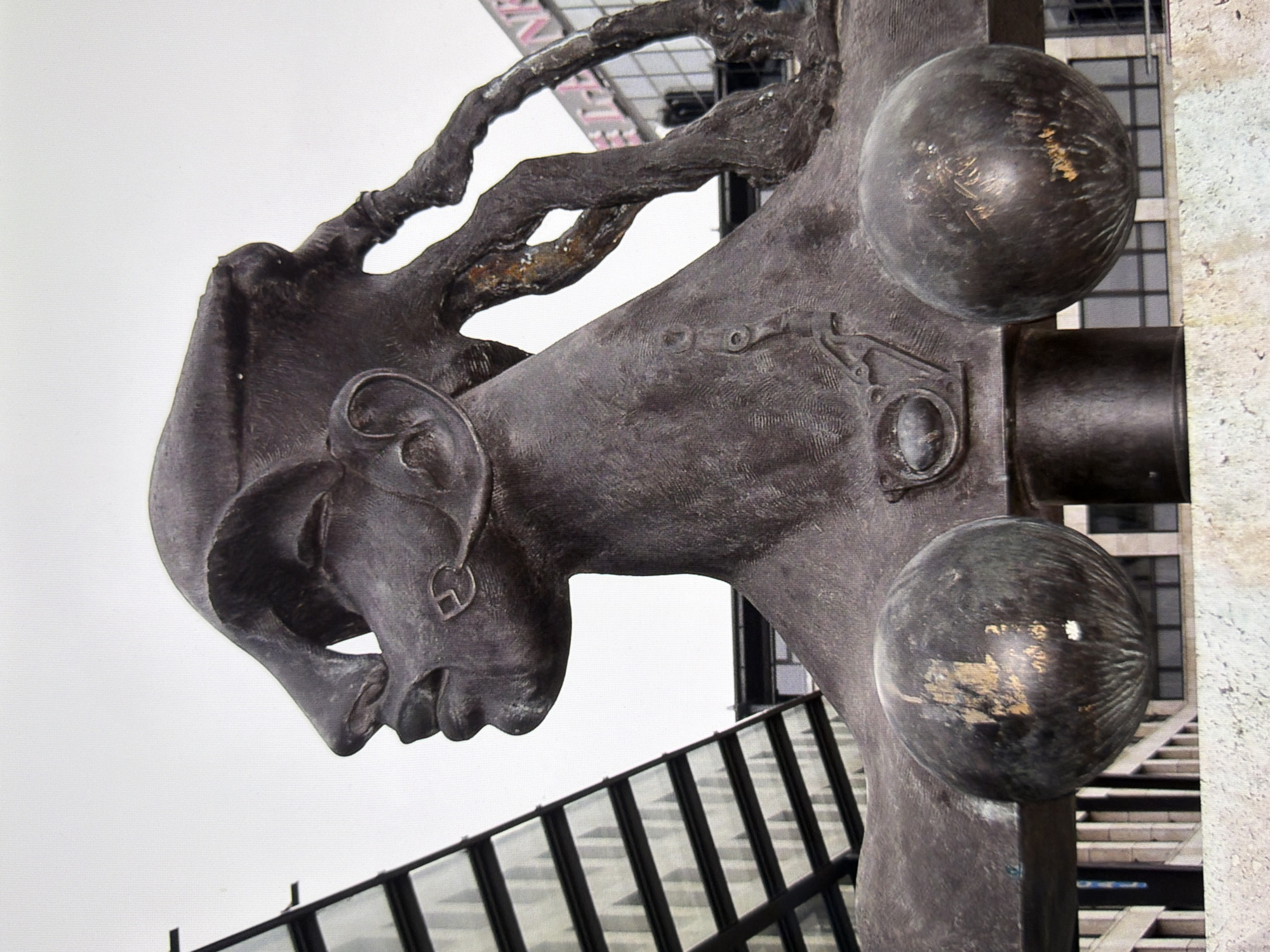 The image shows a close-up view of a dark bronze sculpture depicting the head and upper torso of a stylized human figure. The sculpture has exaggerated facial features, including a prominent, elongated nose and large, rounded lips. The figure wears a helmet-like headpiece that merges into what appears to be flowing hair or tendrils. The sculpture is partially obscured by a modern glass-and-metal building with a diagonal facade, and a portion of another building with pink lettering that is partially visible in the background. The sculpture itself has two large, spherical forms resembling breasts. The base of the sculpture is a cylindrical element with a small, oval emblem. The sculpture appears to be located in an urban environment, possibly a public square or plaza. The location is the "Bronze Woman" sculpture on Maidan Nezalezhnosti (Independence Square) in Kyiv, Ukraine.
