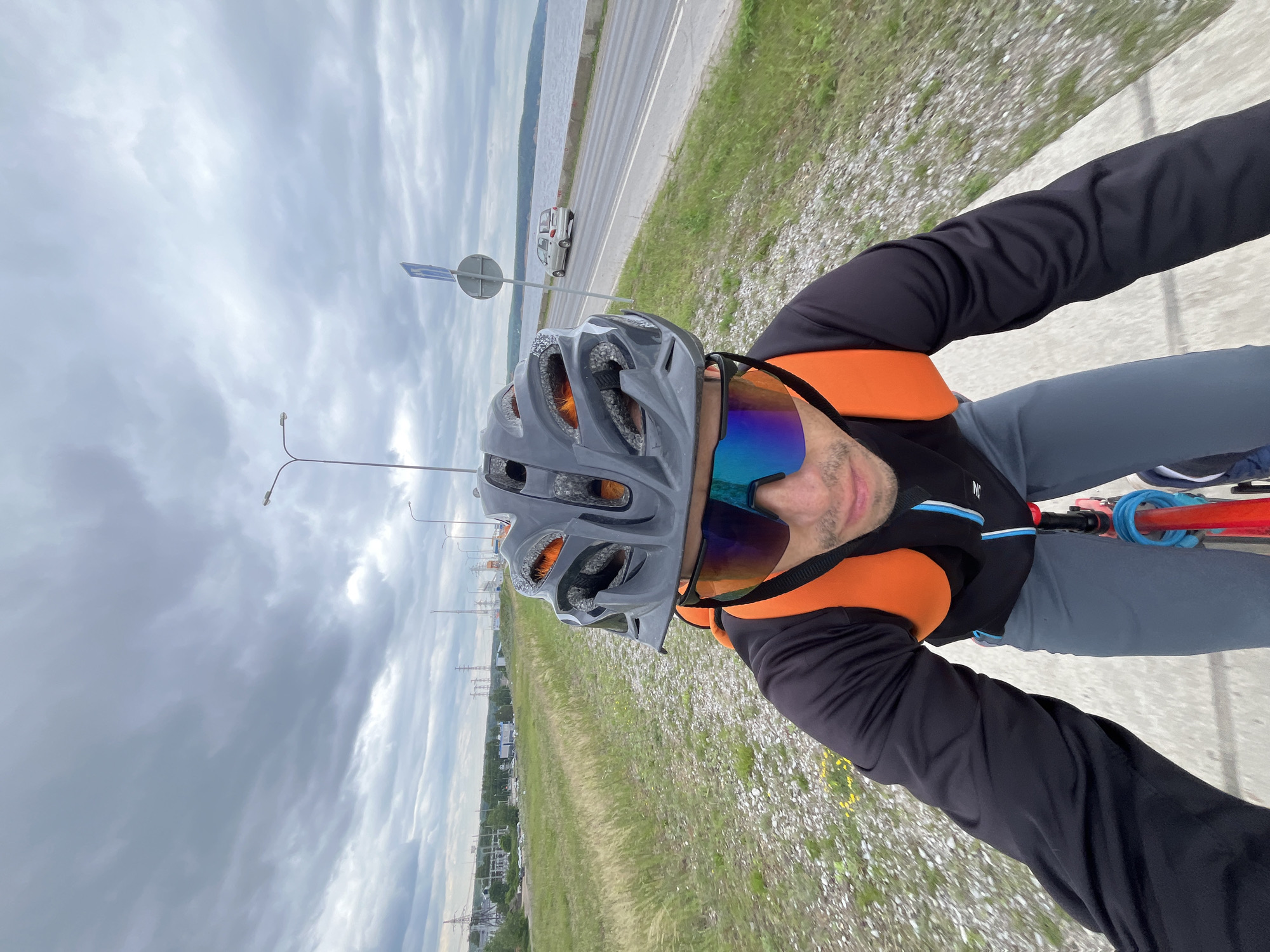 A first-person perspective shot captures a male cyclist wearing a black long-sleeved shirt, grey cycling shorts, a grey helmet with orange accents, and mirrored cycling glasses. He has an orange backpack on and is riding a red bicycle on a paved path. The background features a road with a silver car, a body of water, a grassy embankment, and power lines with electrical towers. The sky is overcast with dramatic clouds. The scene is identifiable as the embankment of the Kama River in the city of Chaikovsky, Russia, a popular location for cycling and recreation.