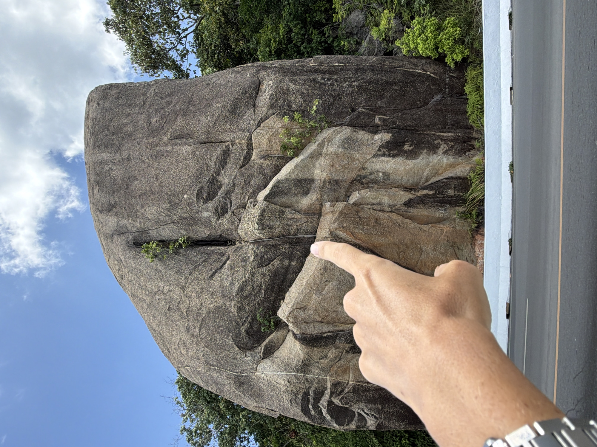 A person's hand, wearing a watch on the left wrist, points towards a large, dark gray boulder that dominates the frame. The boulder is textured with various shades of gray and brown, and shows signs of weathering and cracks. Small green plants sprout from crevices in the rock. The boulder is situated in a lush, green environment with trees and vegetation visible behind it. In the foreground, a gray asphalt road is visible, with a white barrier separating it from the boulder and vegetation. The sky above is blue with scattered white clouds. The location appears to be in Ko Samui District, Thailand.