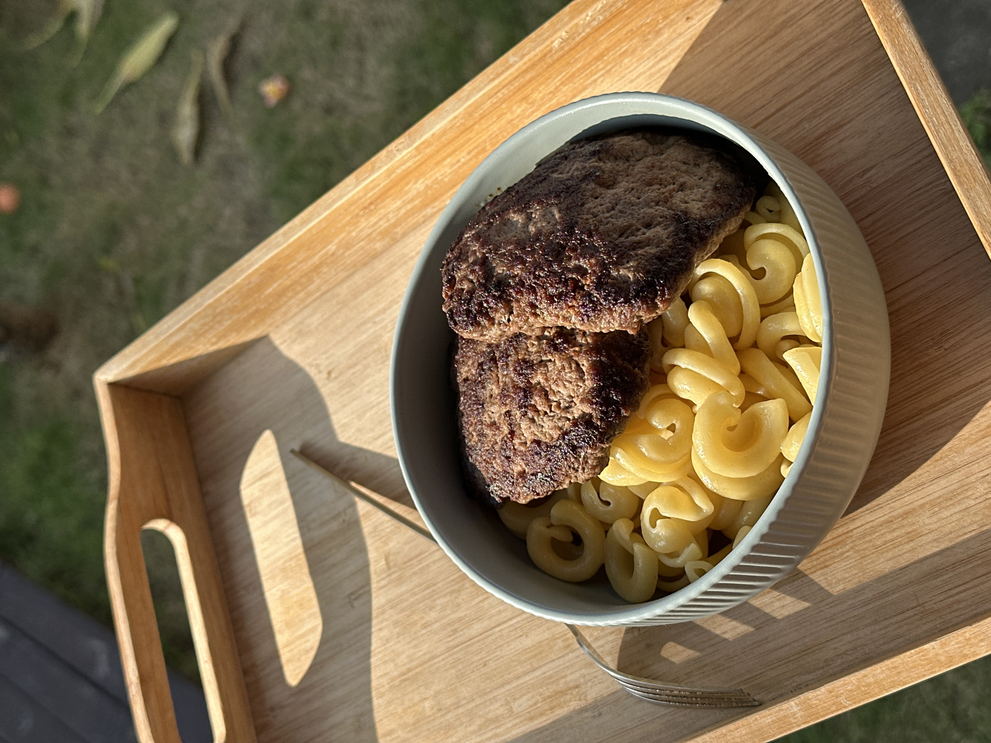 A high-angle shot shows a bowl of pasta with two burger patties on a light-colored wooden tray, presumably outdoors. The location is North Kuta, Indonesia. The bowl is light gray or light blue and contains corkscrew pasta (cavatappi) and two cooked burger patties. The tray is a rectangular wooden serving tray with cut-out handles. A fork rests on the tray beside the bowl. In the blurred background, there is vegetation and some fallen leaves. The lighting suggests a sunny day.