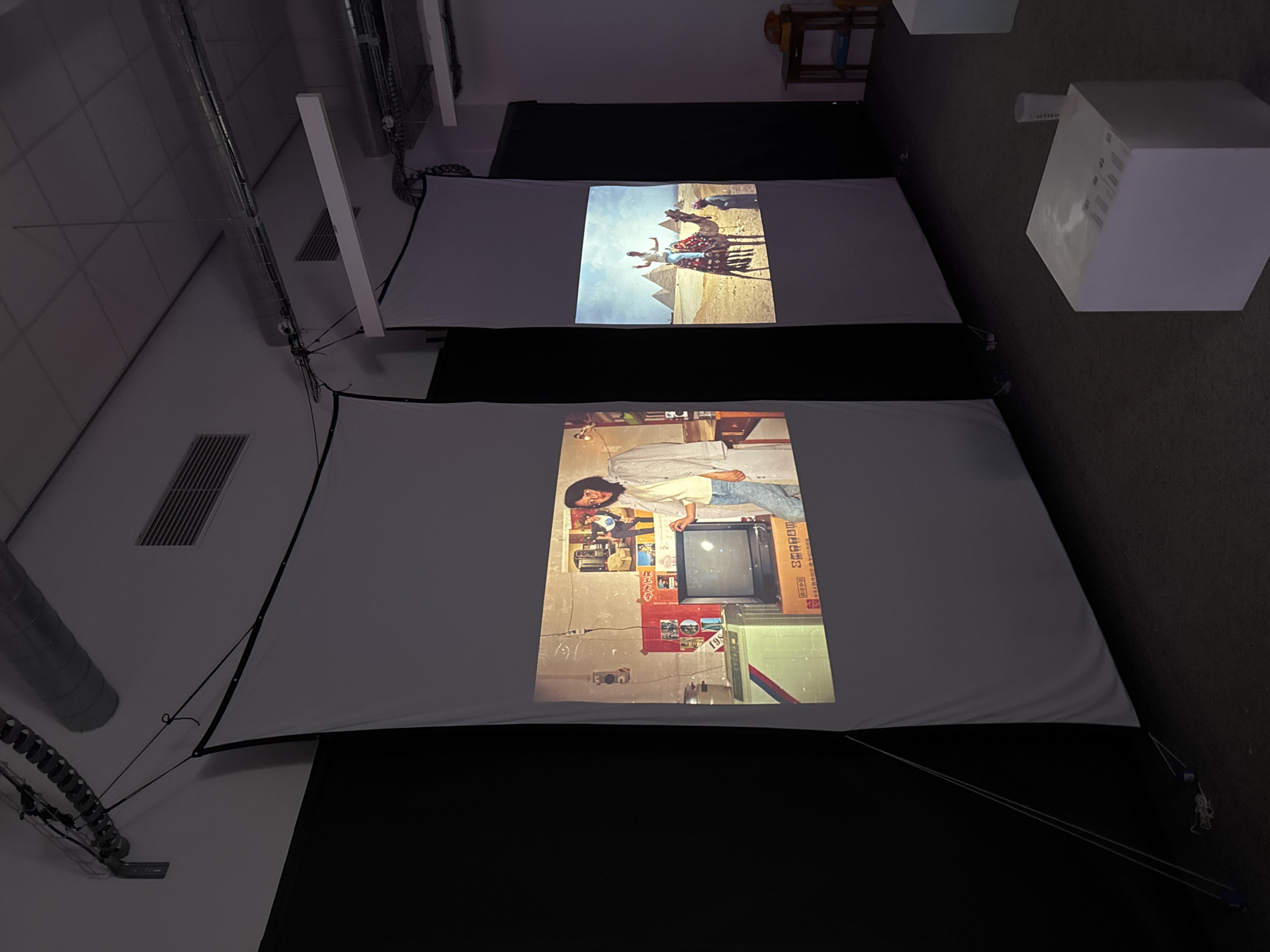 The image depicts an indoor exhibition or viewing space, captured from a high, rotated angle looking down into the room. Two large projection screens are prominently featured, displaying distinct images.
The upper screen projects a vibrant desert scene under a light blue sky. Two individuals are shown riding a camel, with the iconic Giza pyramids visible in the background, suggesting a location in Egypt.
The lower screen displays an interior setting. A person with dark hair, wearing a light-colored long-sleeved shirt and blue jeans, stands or leans next to an older model television set. The wall behind the person and television is covered with various posters or pictures, some with indistinct text. A brown cardboard box next to the television appears to have some printed text or branding, possibly "Panasonic" or similar, though largely unreadable.
The room's ceiling features white square tiles and exposed silver ventilation ducts. White rectangular light fixtures are also visible on the ceiling. Black fabric or panels are used behind and between the screens. In the mid-right background, several white cubic structures are arranged on the floor, some showing faint, unreadable text on their surfaces. A wooden chair or stool is partially visible in the upper right corner. The lighting in the room is dim, accentuating the brightness of the projected images. No specific time of day or emotions are discernible from the scene. The context provided indicates the location is Paris, France.