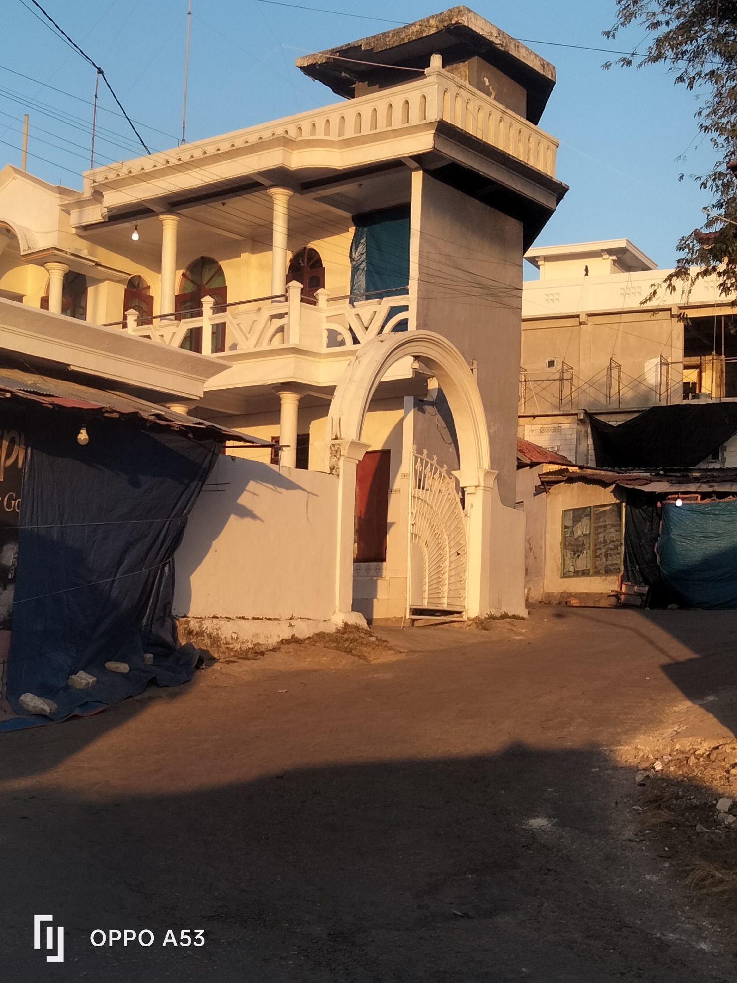 In the city of Badung, Indonesia, a two-story building with white decorative elements and balconies stands under a clear blue sky. The building features arched windows and is partially covered by tarps. In front of the building, there's a pathway leading to a white arched gate with a decorative metal fence. Adjacent structures, some under construction with scaffolding, are visible. The ground is a mix of dirt and asphalt, with shadows cast by the structures and trees. The overall scene depicts a residential or commercial area, likely in a tropical climate given the lighting and vegetation.