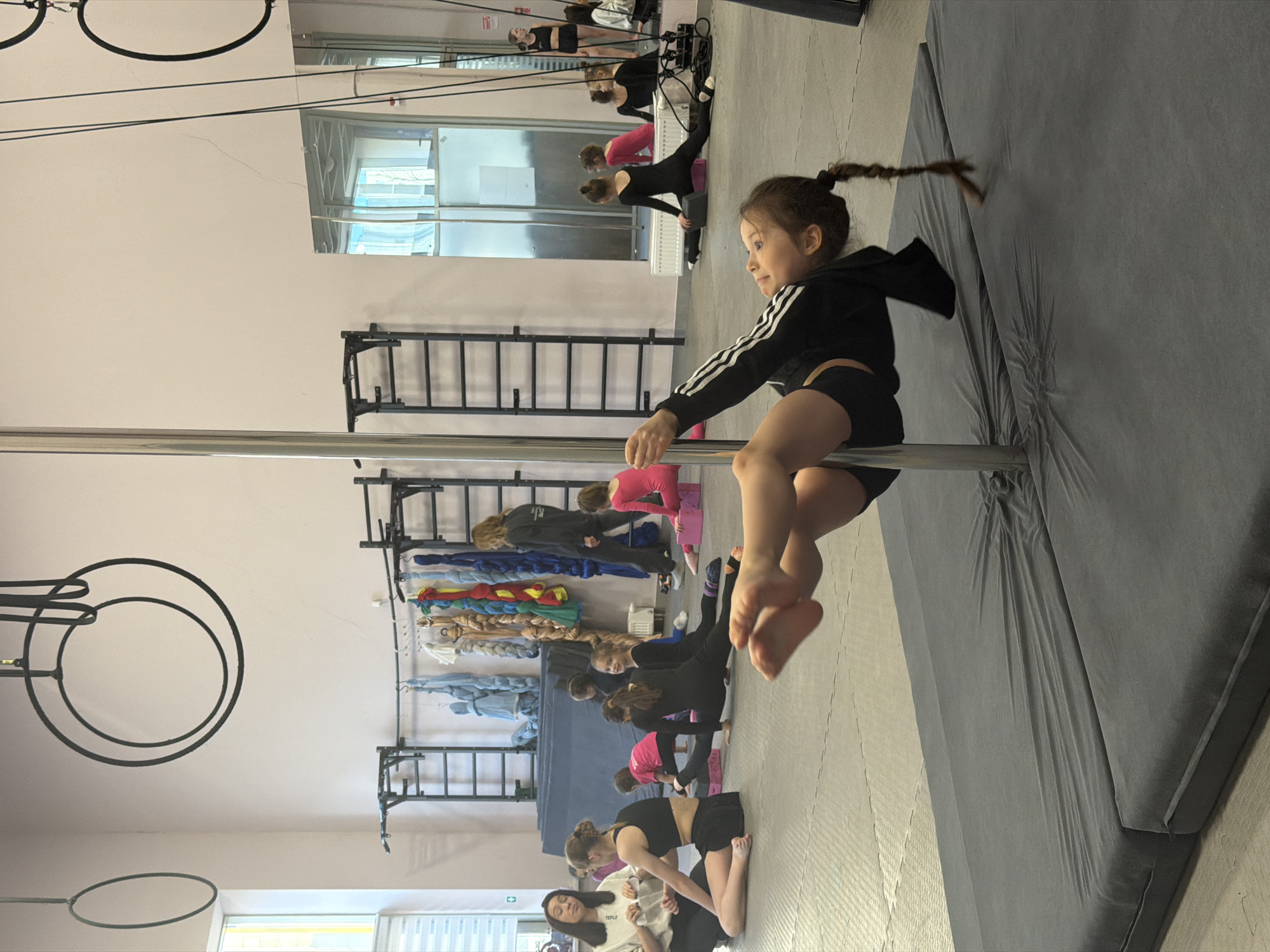 A young girl is shown performing on a pole in what appears to be a dance or fitness studio. She is wearing a black jacket with white stripes on the arms and black shorts. Her hair is pulled back in a braid. A safety mat is beneath her.
In the background, other people are seen stretching and exercising on mats. There is a climbing wall and a rack with clothes. The studio is brightly lit and there are mirrors on the wall.
Based on web searches, this photo seems to be taken at a dance studio in Perm, Russia.