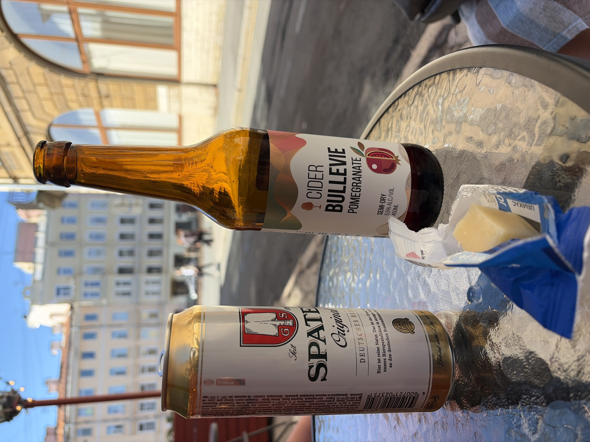In this photograph, taken outdoors in Saint Petersburg, Russia, two beverages are laid out on a glass-topped table. A brown glass bottle of "CIDER BULLEVIE POMEGRANATE" is positioned horizontally, with its label prominently displaying "SEMI-DRY 5.5% ALC/VOL. 450ML". Next to it, a portion of a golden can is visible, featuring the text "SPATE Original DEUTSCHES RE". The can also has some text printed on it in what appears to be Cyrillic script. A small, square piece of cheese, wrapped in white paper and a blue foil, rests on the table near the bottle. The scene is set against a backdrop of blurred buildings and a clear blue sky, suggesting it is daytime and the weather is pleasant. The overall impression is of a casual outdoor setting, perhaps a cafe or a park, where someone is enjoying a drink. The image is oriented vertically, with the bottle and can appearing sideways.
