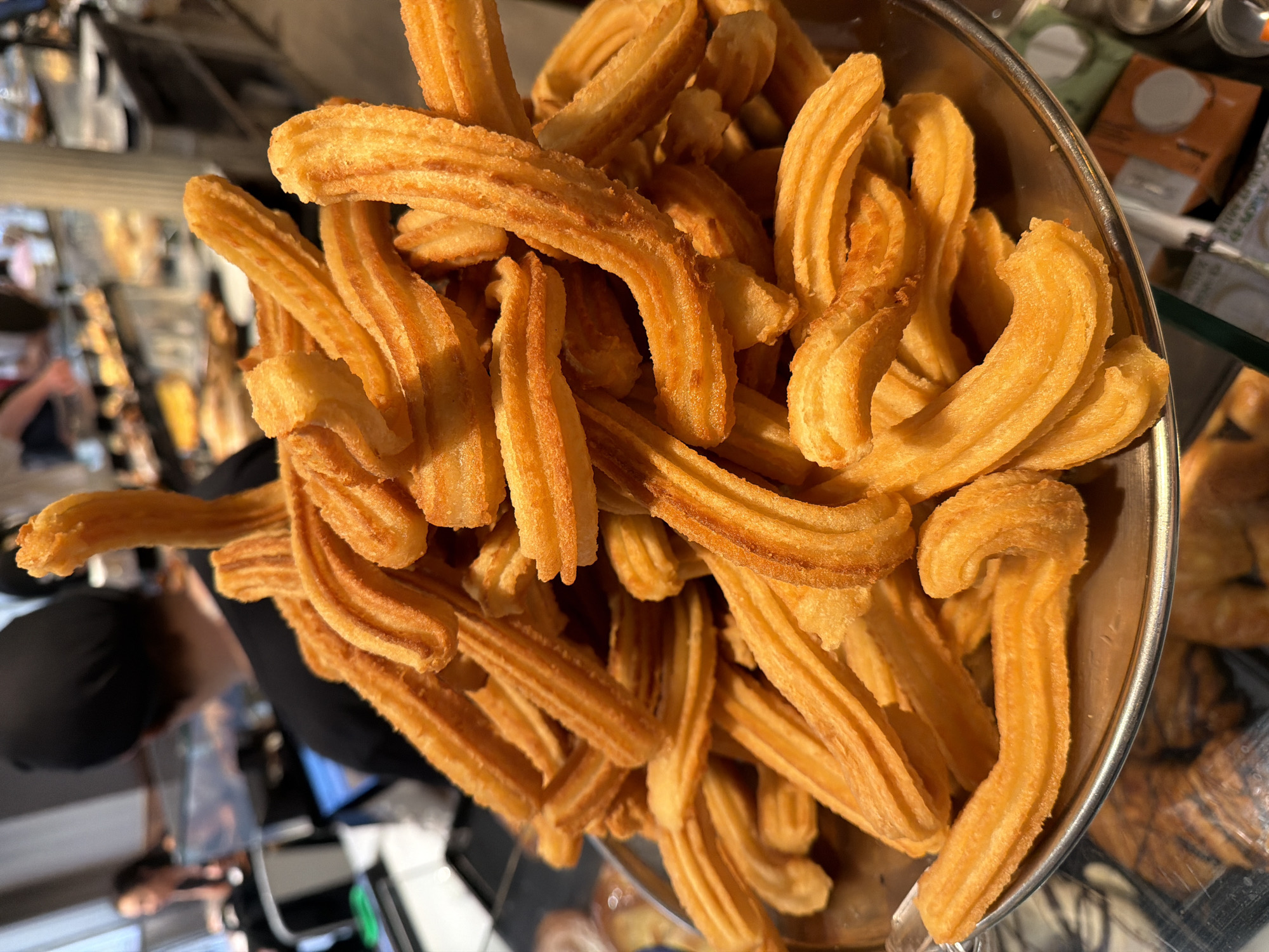 The image captures a close-up, slightly overhead view of a large pile of freshly fried churros on a metallic plate. The churros are golden-brown, ridged, and irregularly shaped, some curved and others relatively straight. They appear to be a popular Spanish pastry, likely from a bakery or a street vendor. In the blurred background, other pastries are visible on shelves, suggesting a food establishment. There are also glimpses of people, with one individual wearing a black hat in focus behind the churros, and another person blurred in the distance. The overall impression is that of a vibrant food scene, possibly in Barcelona, Spain, given the prominence of churros in Spanish cuisine.
