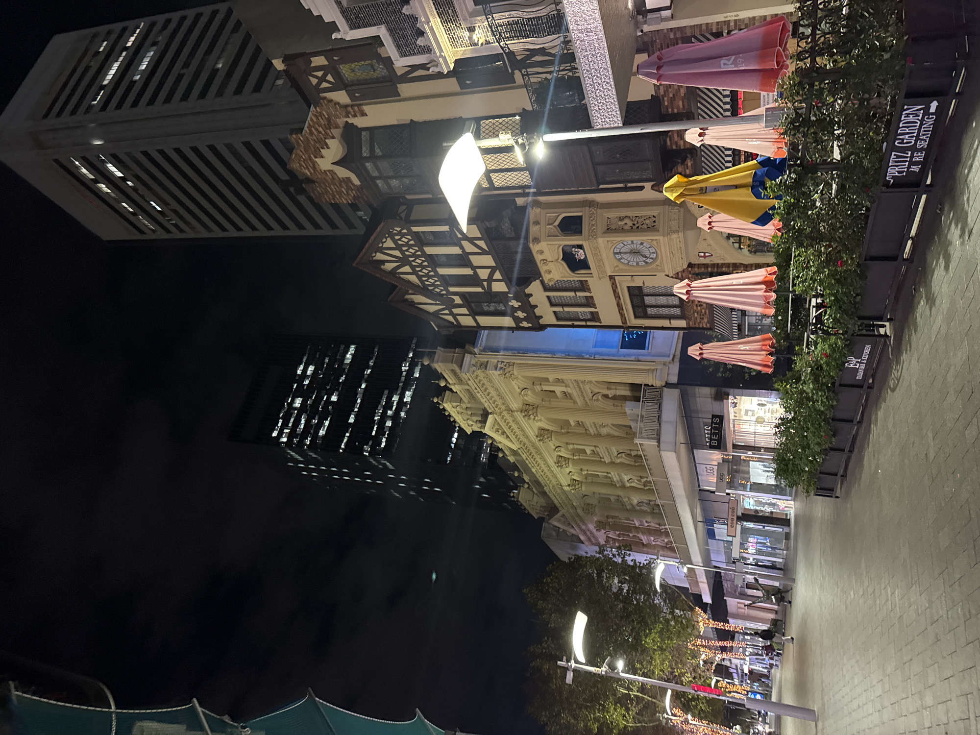 The image shows a street scene at night in Perth, Australia. The camera is at a street-level view pointing skyward, with buildings visible. On the right side of the frame, there's a sidewalk with planters that are labeled, "PRITZ GARDEN M RE SEATING," indicating a garden area next to a restaurant or establishment. A line of closed outdoor umbrellas in pink, yellow, and blue stands behind the planters. The buildings on the right have storefronts with signs such as "TTS BETTS" and various brands like "UGG" visible in the windows.
On the left side of the frame are two prominent buildings. One is a modern skyscraper with many windows reflecting lights. Another is an older building featuring half-timbered architecture and a clock tower. The sky is mostly dark. Street lights illuminate the scene. At the bottom, there is a partial view of what appears to be a tent-like structure with green fabric. In the distance down the street, lights and signage are visible, suggesting commercial activity. People are walking in the distance.
