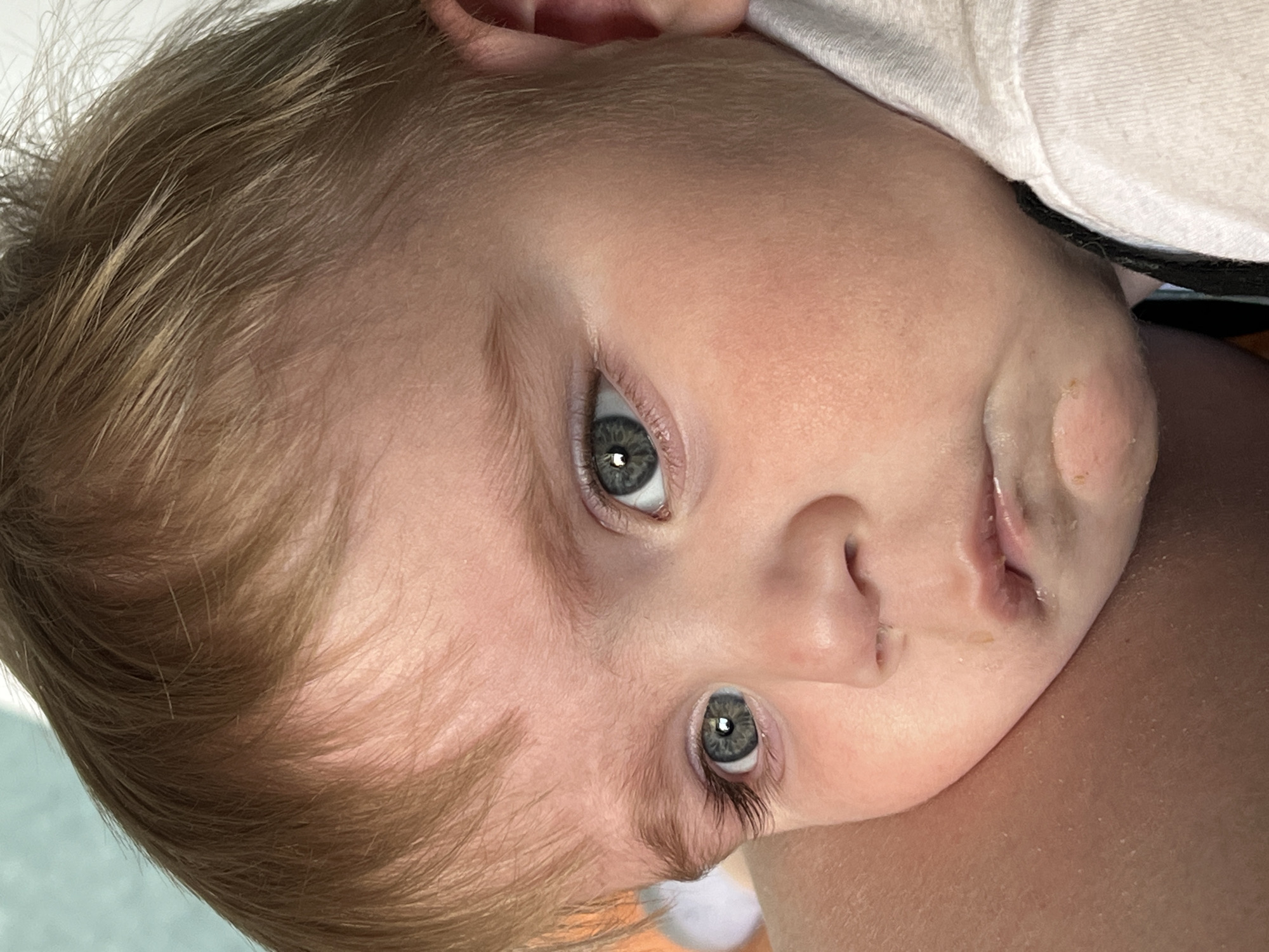 A close-up image shows a baby lying on their side. The baby has blonde hair, pale skin, and grey eyes. The child appears to be resting on someone's shoulder. The background is blurry and indistinct, consisting of light-colored surfaces. There is no discernible information related to the city of Noviy, Russia.