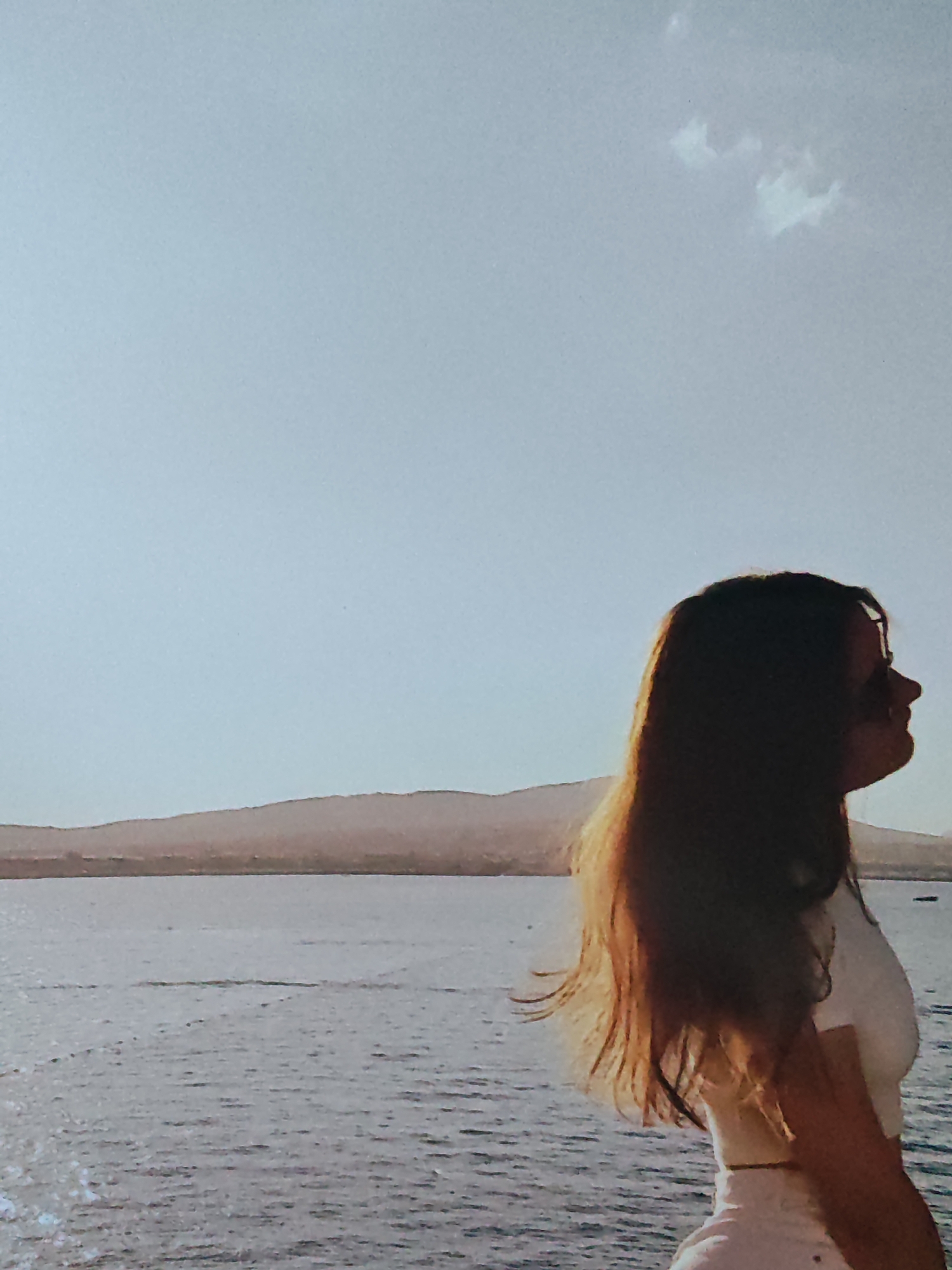 In this outdoor, daytime photograph, a young woman with long, brown hair is captured in profile, facing right. She is wearing a white top and white shorts. The woman stands near the edge of a body of water, which appears to be a lake or the sea. In the background, a hazy, tan-colored mountain range is visible, with a light blue sky above. A small, white cloud is present in the upper right quadrant of the sky. The water's surface is rippled, indicating a gentle breeze. The lighting suggests it is either early morning or late afternoon, with the sun casting a warm glow. The overall scene conveys a sense of tranquility and contemplation.