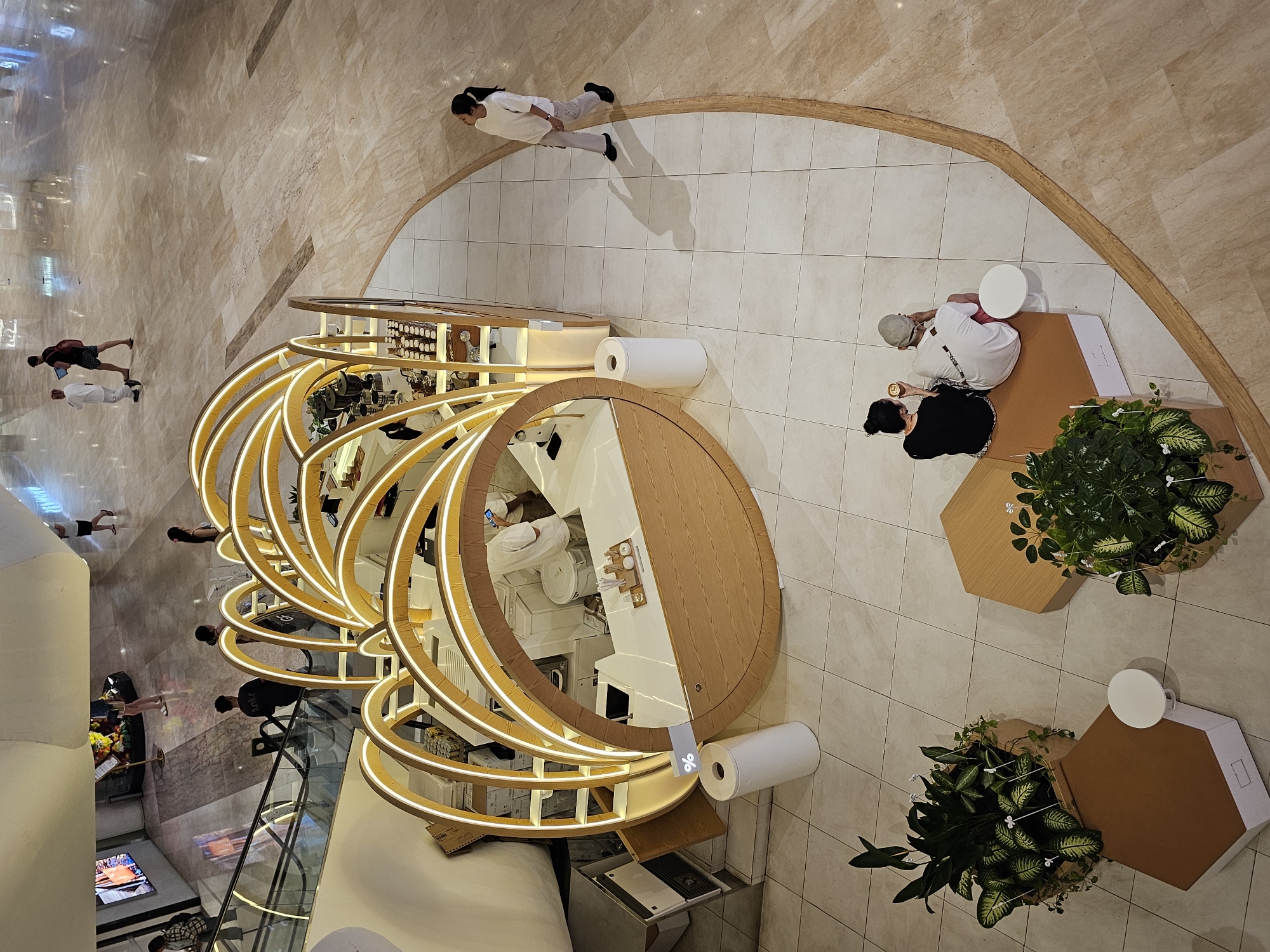 This overhead shot captures a bustling, modern indoor environment, likely a high-end shopping mall or commercial space in Denpasar, Indonesia. The dominant feature is a unique, circular or oval-shaped counter or display unit characterized by multiple golden-hued, illuminated rings that create a striking, layered effect. This counter appears to be a coffee shop or boutique, housing various products and coffee-making equipment visible through its open design.
Several individuals are present, engaged in various activities. One person in a white top and light pants stands near the counter, observing or waiting. Another person in a white shirt, light shorts, and a backpack is seen walking away, holding a blue device. A couple is seated at a small white table next to a hexagonal planter filled with lush green plants. The person in a grey cap, white shirt, and red shorts holds a cup, while their companion, with dark hair, a black top, and a patterned skirt, looks at a phone. Other figures are visible in the distance and reflections, including someone on an escalator, suggesting multiple levels.
The floor is a mix of polished light-colored tiles and larger, irregularly shaped marble or stone sections, contributing to the upscale ambiance. Warm lighting emanates from the counter's rings and overhead fixtures, illuminating the space. A small flag-like sign near the counter displays the distinctive "%" logo, identifying it as an 'Arabica %' coffee shop. The overall atmosphere is clean, contemporary, and relaxed, with people enjoying the amenities of the establishment.