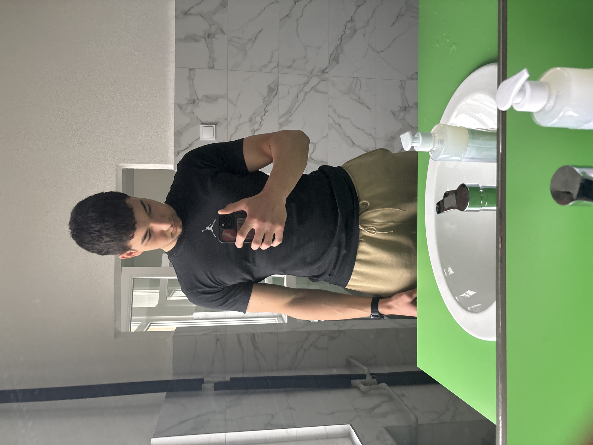 A young man is taking a selfie in a bathroom. He is wearing a black t-shirt with a Jordan logo, and tan sweatpants. He is holding a smartphone in his right hand. The background consists of marble wall tiles and a mirror. A sink with a green cabinet is visible on the right side of the mirror. The location is not explicitly mentioned in the image. Without further information, I cannot confirm that this image was taken in Zlatograd, Bulgaria.