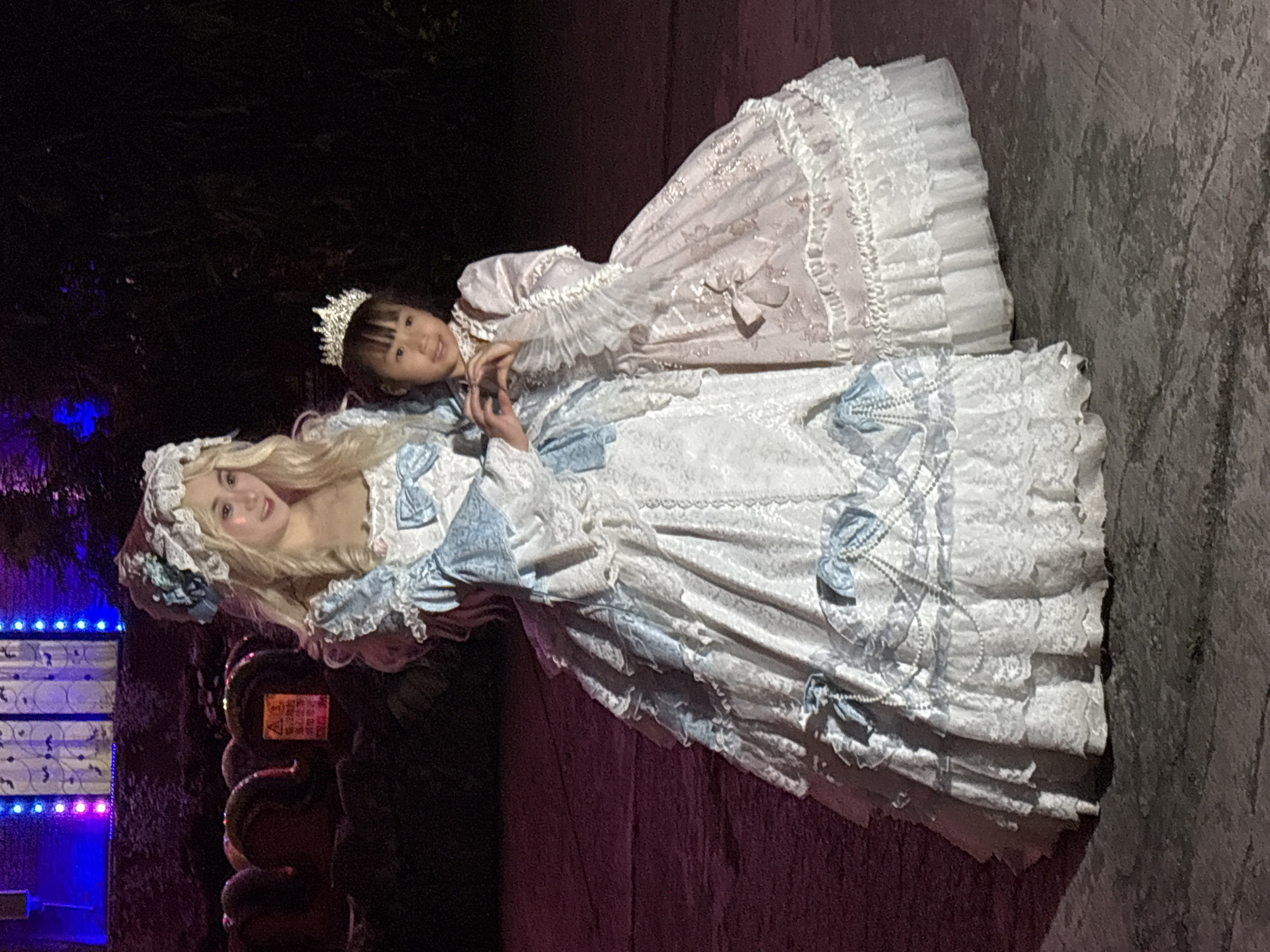 Two individuals are visible in the image, positioned outdoors, likely in the city of Baqiao, China. One is an adult woman, wearing a long, white, layered dress with blue accents and lace. Her hair is blonde, and she wears a white bonnet. The second individual is a young girl wearing a similar, long dress in light pink and white, with a silver tiara on her head. The two are positioned closely, suggesting interaction.
The background includes a wall to the right, and to the left, an illuminated structure with blue and pink lights is visible. The lighting conditions suggest it might be nighttime.
