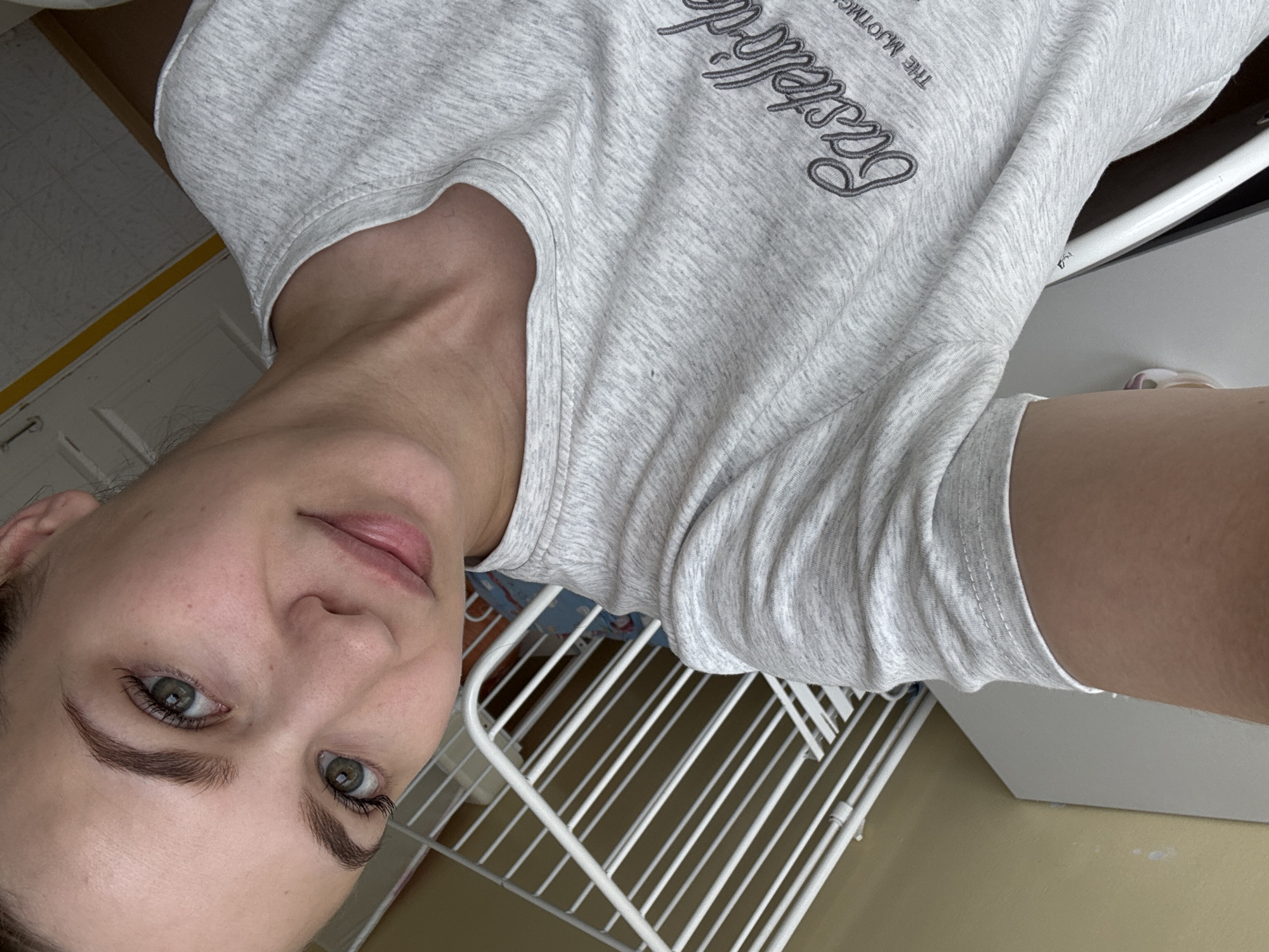 A frontal, waist-up, slightly tilted selfie-style photograph depicts an adult female indoors in Chaikovsky, Russia. She has fair skin, well-defined dark eyebrows, and green eyes with prominent eyelashes. Her lips are naturally colored and slightly parted, indicating a gentle expression. Her dark hair is pulled back from her face.
She is wearing a light grey, heathered short-sleeved t-shirt. Dark grey script text is embroidered or printed on the chest, partially legible as "Castelló de" followed by a smaller line reading "THE MJOTMG".
The background on the lower left features a white, multi-barred structure, consistent with a crib, child's bed rail, or shelving unit. Behind this structure and extending to the upper left is a plain light yellow-beige painted wall, which exhibits several light-colored smudges or marks. The upper right background reveals a white tiled ceiling, a partial view of a white door, and a thin yellow strip running along the wall-ceiling junction. The lighting is ambient and even, illuminating the scene without harsh shadows, consistent with an indoor residential or domestic environment.