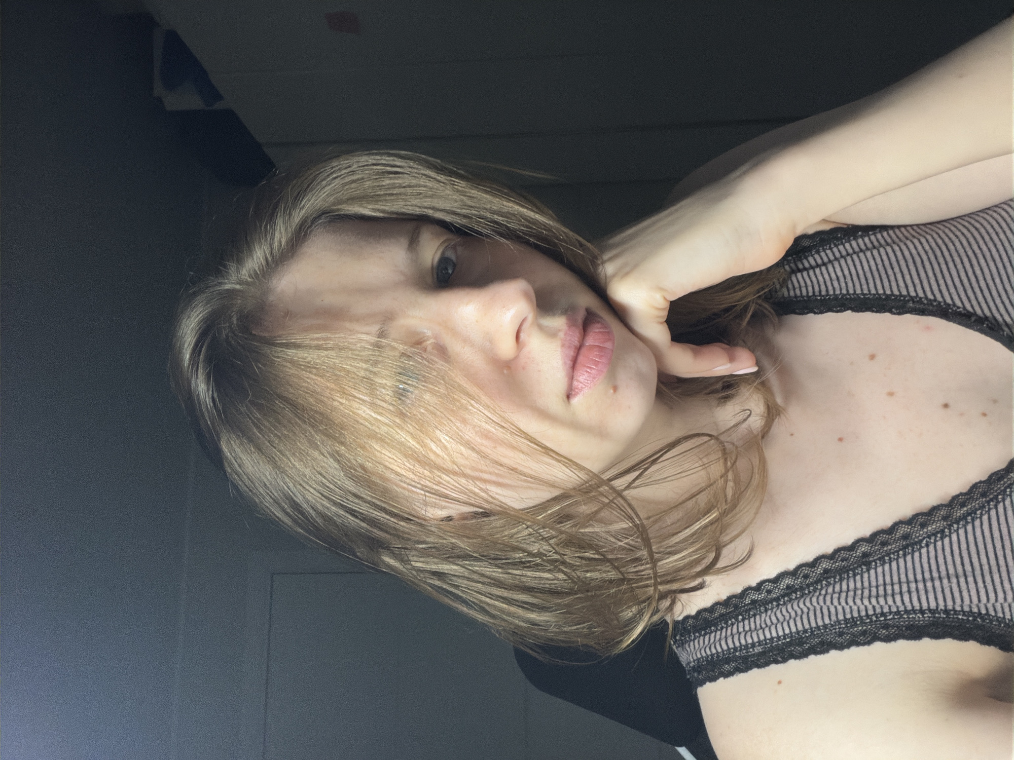 A young woman with light brown hair is shown in a close-up, head-and-shoulders shot. She is looking slightly to her right, with her right hand resting on her chin. Her expression is neutral, and her lips are slightly parted. She is wearing a black and white striped tank top with black lace trim. The background is dark and out of focus, with hints of furniture and a wall. The lighting is dim, with a light source coming from the left, casting shadows on her face. The image appears to be taken indoors, possibly in a bedroom or living area. There is no specific indication of the city of Dongducheon-si, South Korea.