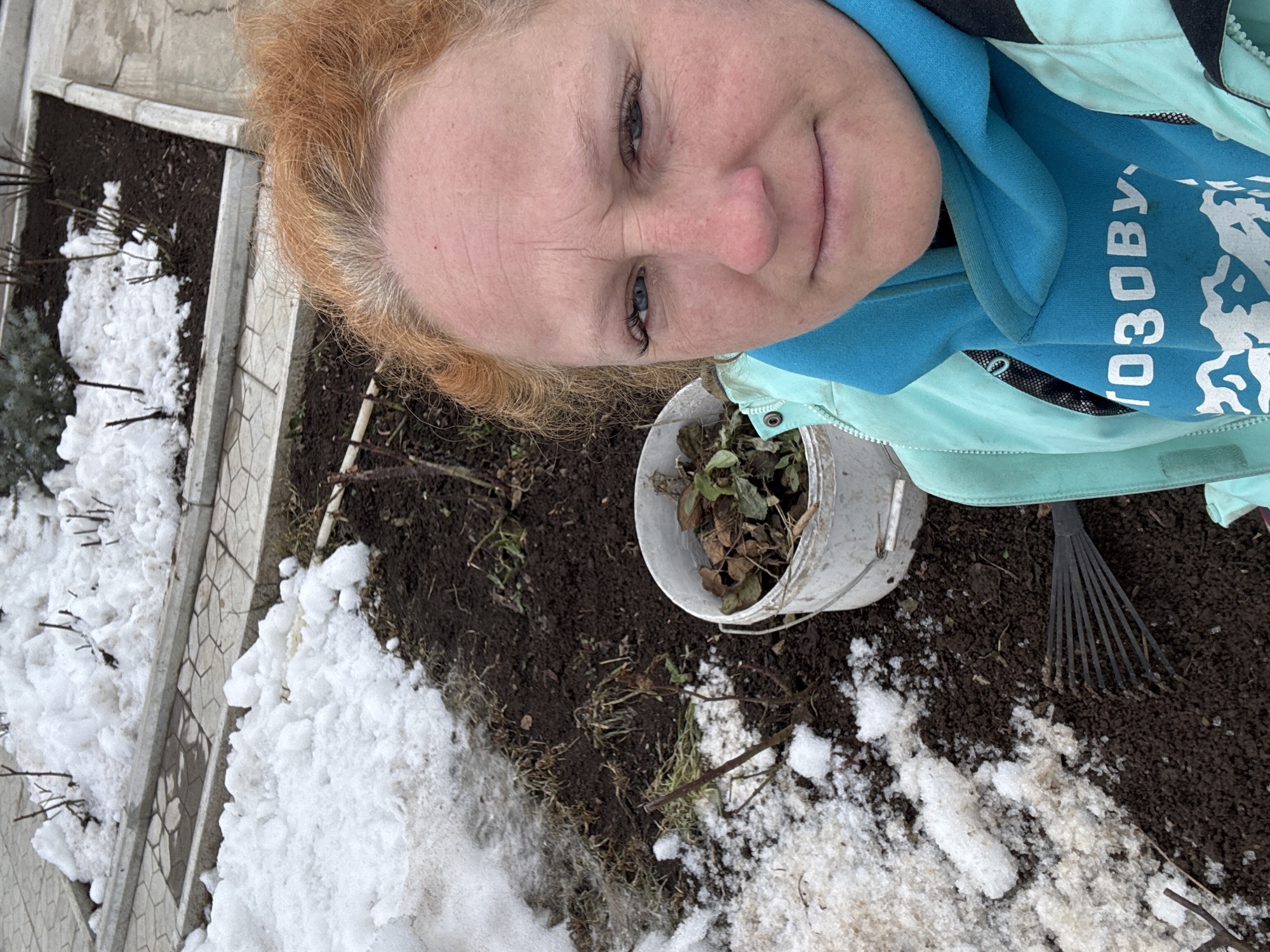 A woman with red hair and blue eyes is taking a selfie outdoors in what appears to be a garden or yard. She is wearing a light blue jacket over a blue shirt with the Russian word "СОЗОВУ" on it. A white bucket containing dead leaves is visible. There is a small rake on the ground. The ground is covered in dirt and patches of snow. There are also plant beds with some snow present. Based on the language visible on the shirt, and potentially snowy conditions on the ground, it is consistent with the location of the city of Chaikovsky, Russia.