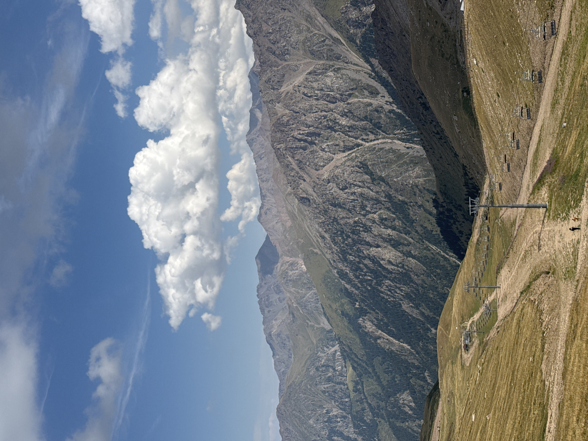 A wide, high-angle shot captures a vast mountainous landscape under a partly cloudy blue sky. In the foreground, a dry, grassy slope features a dirt path winding its way up. A ski lift with multiple empty chairs stretches across the midground, ascending the slope. Two small figures walk along the path in the lower right corner. Behind the grassy slope, a series of rugged, rocky mountains rise, their slopes partially covered in dark green vegetation. The sky is dotted with fluffy white clouds. The scene appears to be a ski resort in a mountainous region during the daytime.