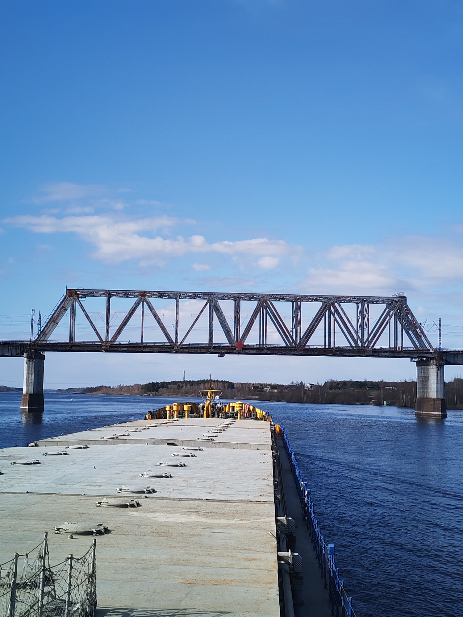 This media file captures a wide river scene under a clear, bright blue sky with scattered white clouds. The perspective is from the deck of a large, flat-bottomed vessel, possibly a barge, looking forward.
In the foreground, the expansive, light-grey deck of the vessel is prominent, featuring numerous tie-down points or hatches. A blue railing runs along the starboard side of the vessel, enclosing what appears to be a walkway. Towards the front right of the deck, some yellow machinery or equipment is visible. The vessel appears to be moving, indicated by the ripples and wake in the dark blue water of the river alongside its hull.
Dominating the midground is a sturdy metal truss bridge, likely a railway bridge, spanning the wide river. The bridge structure is a classic lattice design, painted a reddish-brown and supported by robust concrete piers on either bank and within the water. Railway tracks and overhead electrification lines are visible on the bridge deck. On the truss structure itself, a white sign with the number "17" is faintly discernible on the right side.
The river extends into the distance, flanked by tree-lined shores on both sides. Scattered buildings can be seen amidst the trees on the distant land. The overall impression is one of calm, clear weather, likely during the day, as the sun illuminates the scene and reflects off the water. The location context indicates this scene is in Tarkanovo, Russia.