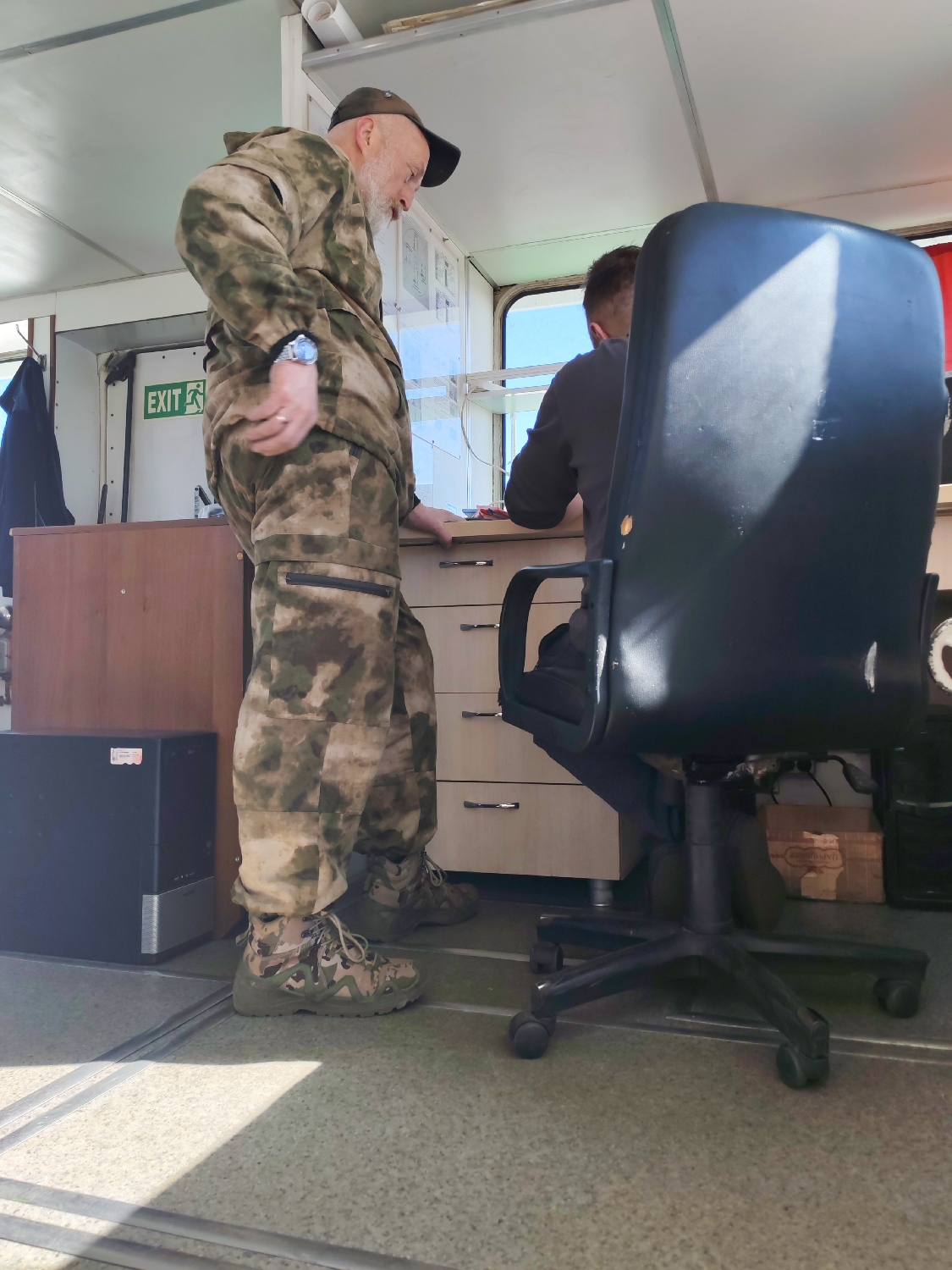 This image captures an indoor scene, likely a functional space such as a control room, cabin, or office on a vessel, given the window structure and utilitarian interior design. Two men are present, illuminated by bright daylight streaming in from a large window.
In the foreground, an older man with a white beard and a dark baseball cap stands, facing towards the right side of the frame. He is dressed in a full camouflage uniform, including a jacket and pants in shades of green, brown, and beige, with tactical-style boots on his feet. A wristwatch is visible on his left wrist, and he wears a ring on his left hand. His posture suggests he is either observing something or engaged in conversation with the other person.
To the right, partially obscured by a large, black office chair on wheels, sits a younger man. Only his back and the back of his head are visible, as he faces away from the viewer, appearing to be focused on a light-colored desk or console in front of him.
The room features light-colored walls and ceiling, with what appears to be metal framing or trim. A prominent green "EXIT" sign is affixed to a wall to the left of the older man. On the floor, a black rectangular object, possibly a computer tower or speaker, sits next to the older man's feet. The floor is light-colored and textured. Strong shadows are cast across the floor and the office chair, indicating direct sunlight from the window. The sky visible through the window is bright blue, suggesting clear weather. Underneath the desk, various items, including a cardboard box, are visible.