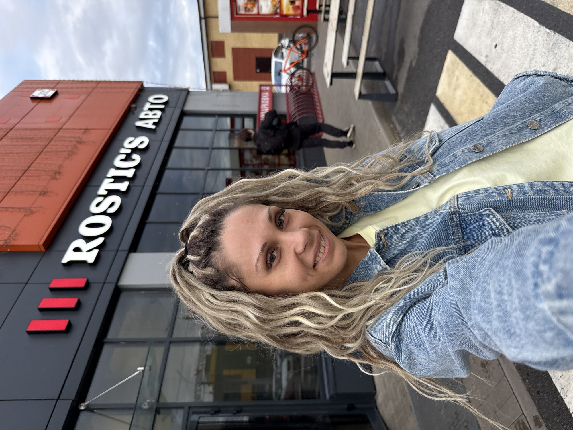 A woman with blonde hair tied in a ponytail and wearing a denim jacket and a yellow shirt takes a selfie in front of a "ROSTIC'S ABTO" building in Ignatovo, Russia. A man in a black jacket and backpack walks past a bicycle rack and a white car is parked in the background. The sky is overcast. There are picnic tables and benches in the foreground.