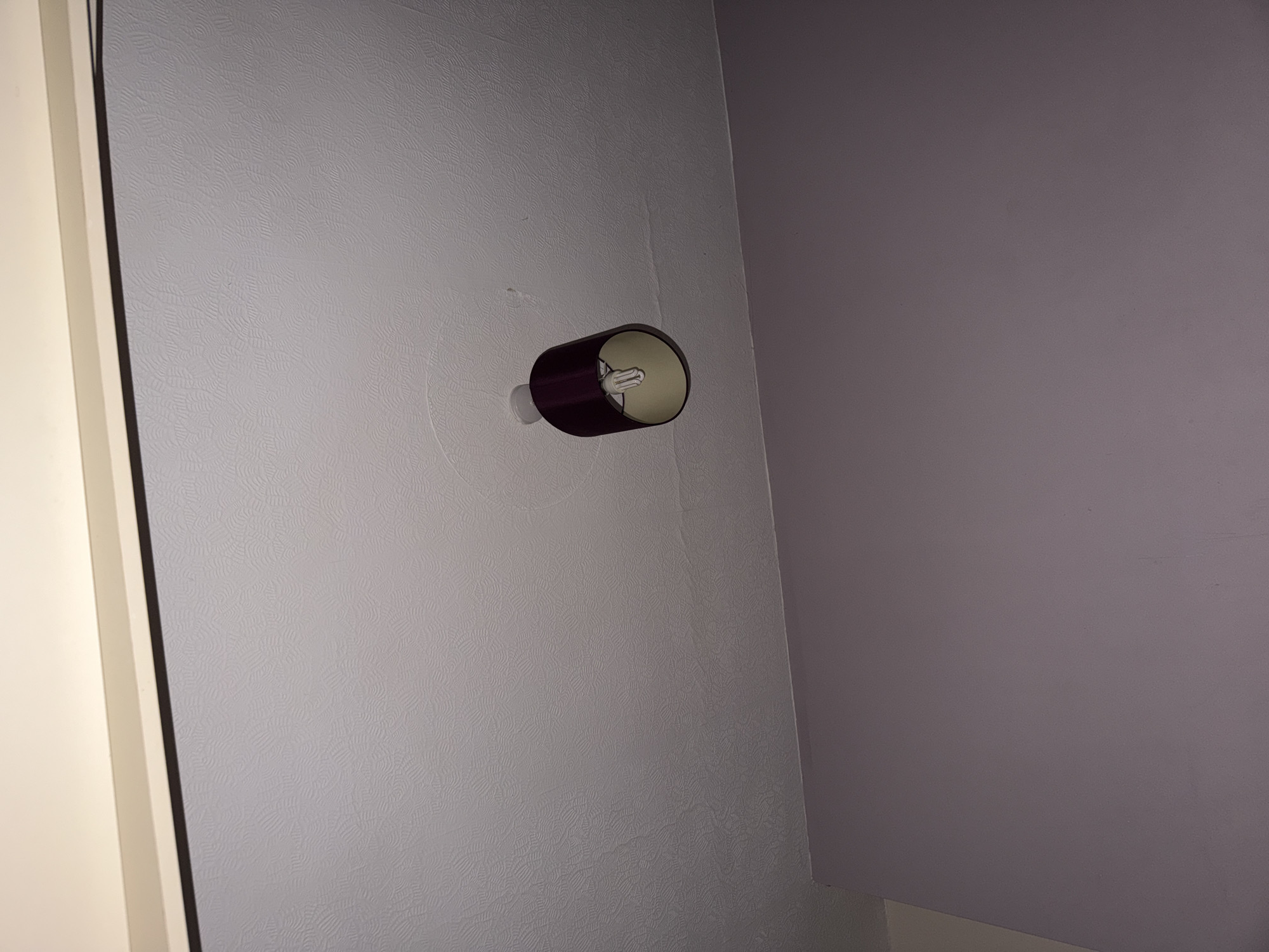 The image shows an indoor scene, likely a residential interior, with a light fixture mounted on a wall. The fixture has a burgundy shade, cylindrical in shape, and appears to be a sconce type. A lightbulb is visible within the shade. The wall it's mounted on is textured and painted in a light greyish-white hue. Adjacent to this wall is another one painted a darker shade of greyish-purple. The corner where the two walls meet is also visible. To the left of the frame is a portion of a door frame, painted white. No people or other objects of significance are present. The image does not provide enough information to determine the specific location within Cwmbrân, United Kingdom.