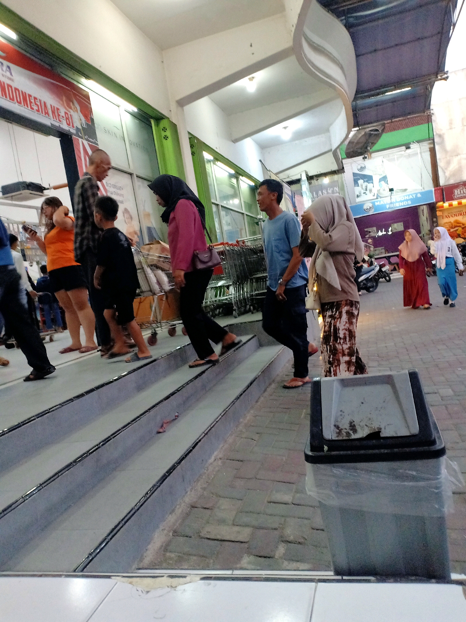 The image captures a scene in Bojonegoro, Indonesia, likely a shopping area or a public walkway. Several individuals are visible, walking on a sloped surface that resembles an outdoor escalator or a series of ramps. In the foreground, a grey trash bin with a plastic liner is prominent.
To the left, a group of people are walking up the incline. A woman in an orange shirt and dark shorts is looking at her phone. Next to her, a man in a plaid shirt is walking, and a young boy in a black shirt and shorts is following him. Further left, out of focus, are more people and shopping carts.
In the center and right of the image, other individuals are also moving along the walkway. A man in a blue shirt and dark pants walks past. A woman in a tan hijab and a patterned skirt is also present. Further to the right, two women are walking away from the viewer; one in a long red dress and the other in a light blue hijab and white jacket. In the background, there are store fronts and signs, including one with "MAYO DONAT & FRIENDS" and another with "FAVOURI..." possibly advertising food. A motorcycle is parked on the right side of the walkway. The overall environment appears to be a covered outdoor area, with a ceiling structure visible at the top of the image and green painted structural elements.