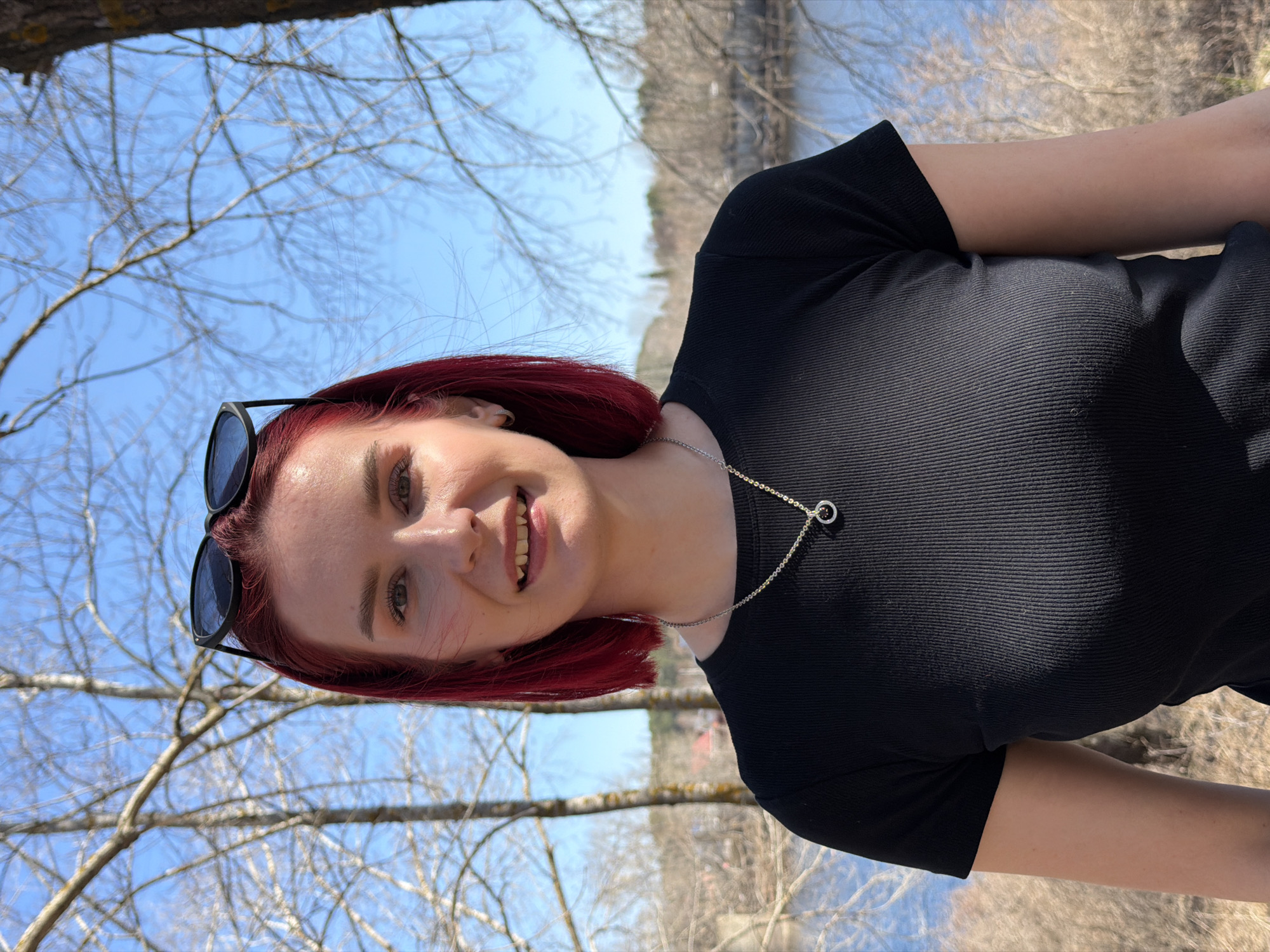 A young woman with red hair stands outdoors, smiling. She wears a black top and a silver necklace with a ring pendant. Sunglasses are perched on her head. The background features a blue sky, bare trees, and a glimpse of water. The location is Chaikovsky, Russia.