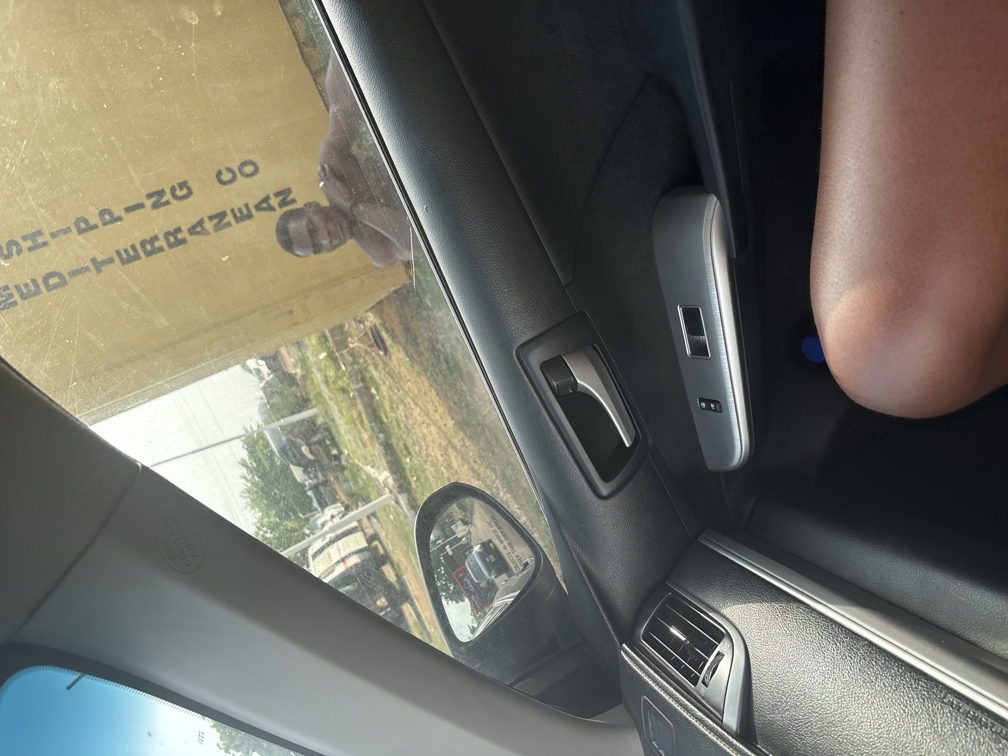 The image, taken from inside a vehicle, captures a view through the passenger side window, rotated to appear upright. On the right, a person's light brown-skinned arm or upper thigh is visible, indicating a passenger. The car's interior shows a black textured door panel with silver trim, a silver door handle, and window control buttons. An air vent is visible towards the bottom right. Above the windshield, an "SRS CURTAIN AIRBAG" label is present on the A-pillar. The side mirror shows the usual warning text "OBJECTS IN MIRROR ARE CLOSER THAN THEY APPEAR" and reflects other vehicles.
Through the passenger window, a large yellowish surface is visible, bearing the prominent text "MEDITERRANEAN SHIPPING CO". This suggests the vehicle is parked near or passing by a commercial building, shipping container, or large sign. Beyond this reflection, an outdoor scene unfolds with green trees and what appears to be a road or open area with other vehicles in the distance. The bright lighting indicates it is daytime and sunny. The overall scene suggests a setting in a commercial or industrial area, possibly a port or logistics hub, consistent with the provided location context of Iddo Tudunwada, Nigeria.