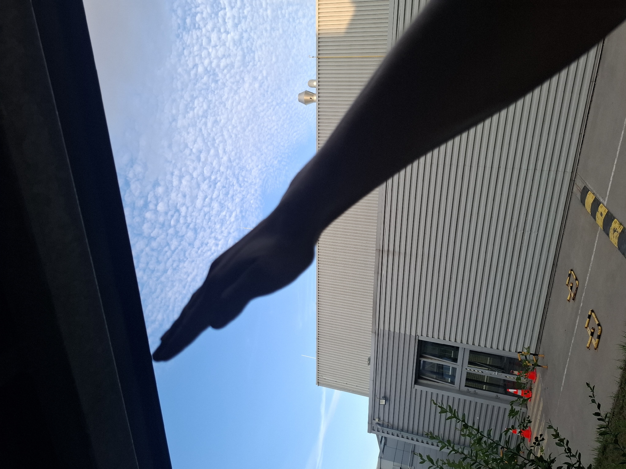 The image, taken from a low angle, shows the silhouette of a person's arm and hand extended upwards against a bright blue sky filled with scattered, puffy white clouds. The hand is held flat, with fingers together, as if making a gesture. Below the sky, a corrugated metal building is visible, with several industrial-looking vents or structures on its roof. In the foreground, there are orange traffic cones and what appear to be speed bumps on an asphalt surface. The overall scene suggests an outdoor, possibly industrial or commercial area, bathed in daylight. The image was captured in Sedlec, Czechia.