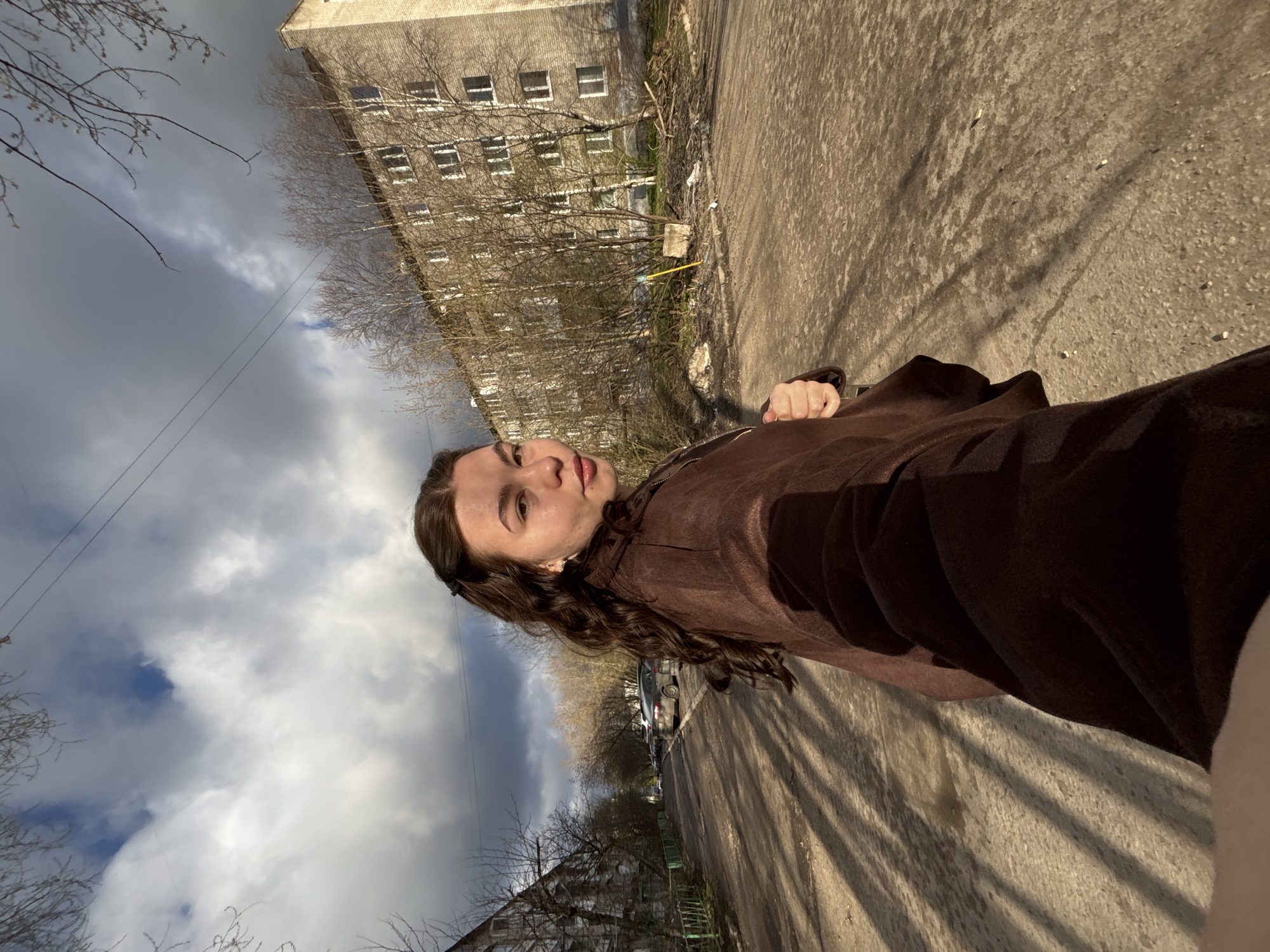 The image captures a young woman posing for a photo outdoors in an urban setting, likely a street or pathway, with the entire scene rotated 90 degrees clockwise. The main subject is a young woman with long, dark, wavy hair, wearing a brown zippered jacket. She looks directly towards the camera with a neutral expression.
The background features multi-story, greyish-brown brick apartment buildings, characteristic of Soviet-era architecture, with numerous rectangular windows. Bare trees line the street in front of the buildings, suggesting late autumn, winter, or early spring. The paved ground, likely asphalt or concrete, shows distinct long shadows cast by the trees, indicating a sunny day, possibly mid-morning or afternoon. Some parked cars are visible in the distance along the street.
The sky is partially cloudy, with varying shades of grey clouds and some patches of brighter, possibly blue, sky visible. Thin power lines cut across the upper portion of the frame. The overall atmosphere appears calm. There is no visible text in the image. The architectural style and general environment are consistent with the given location context of Perm, Russia.