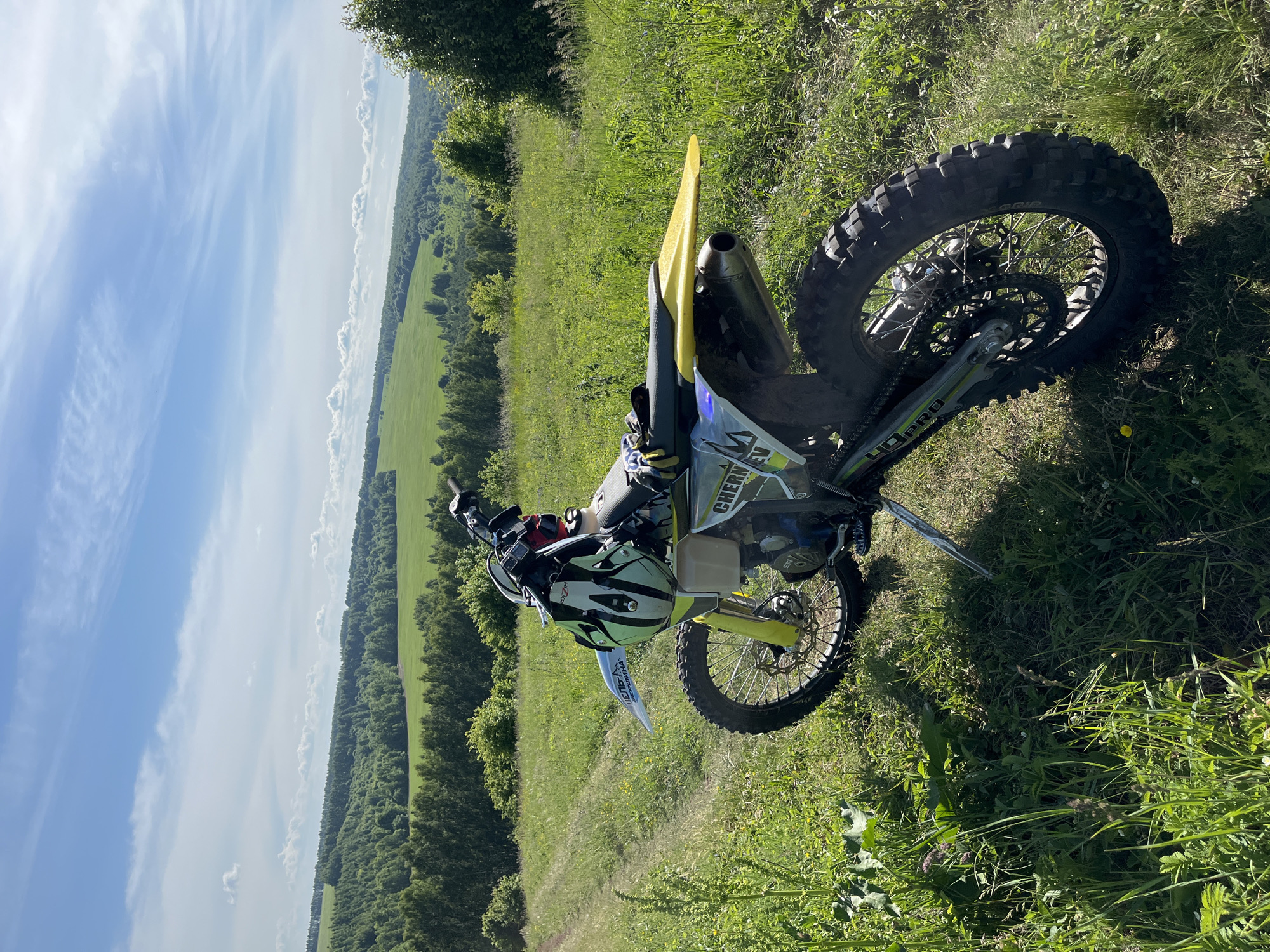 The image displays an off-road motorcycle parked on a grassy incline overlooking a rural landscape in Vorob'evka, Russia. The motorcycle is positioned with its front section towards the left and its rear towards the lower right of the frame, supported by a deployed kickstand. It features a prominent yellow rear fender, yellow front fork guards, and a white front fender with visible Cyrillic text including "ЦЕЛЬ-А" and "РЯБИНА." The side fairing includes the text "CHERN EV." A green, white, and black motocross helmet, along with yellow and blue gloves, rests on the motorcycle's seat or handlebars. The vehicle is equipped with knobby tires suitable for rough terrain.

The foreground consists of lush green grass and various wild plants. A narrow, unpaved track is visible on the left side, curving slightly uphill. The midground and background reveal a vast expanse of rolling hills covered in dense green forests, interspersed with bright green fields, likely agricultural. The sky above is clear blue with scattered white cumulus and cirrus clouds. The scene is illuminated by direct sunlight, casting distinct shadows, suggesting midday or early afternoon. No individuals are visible in the image.