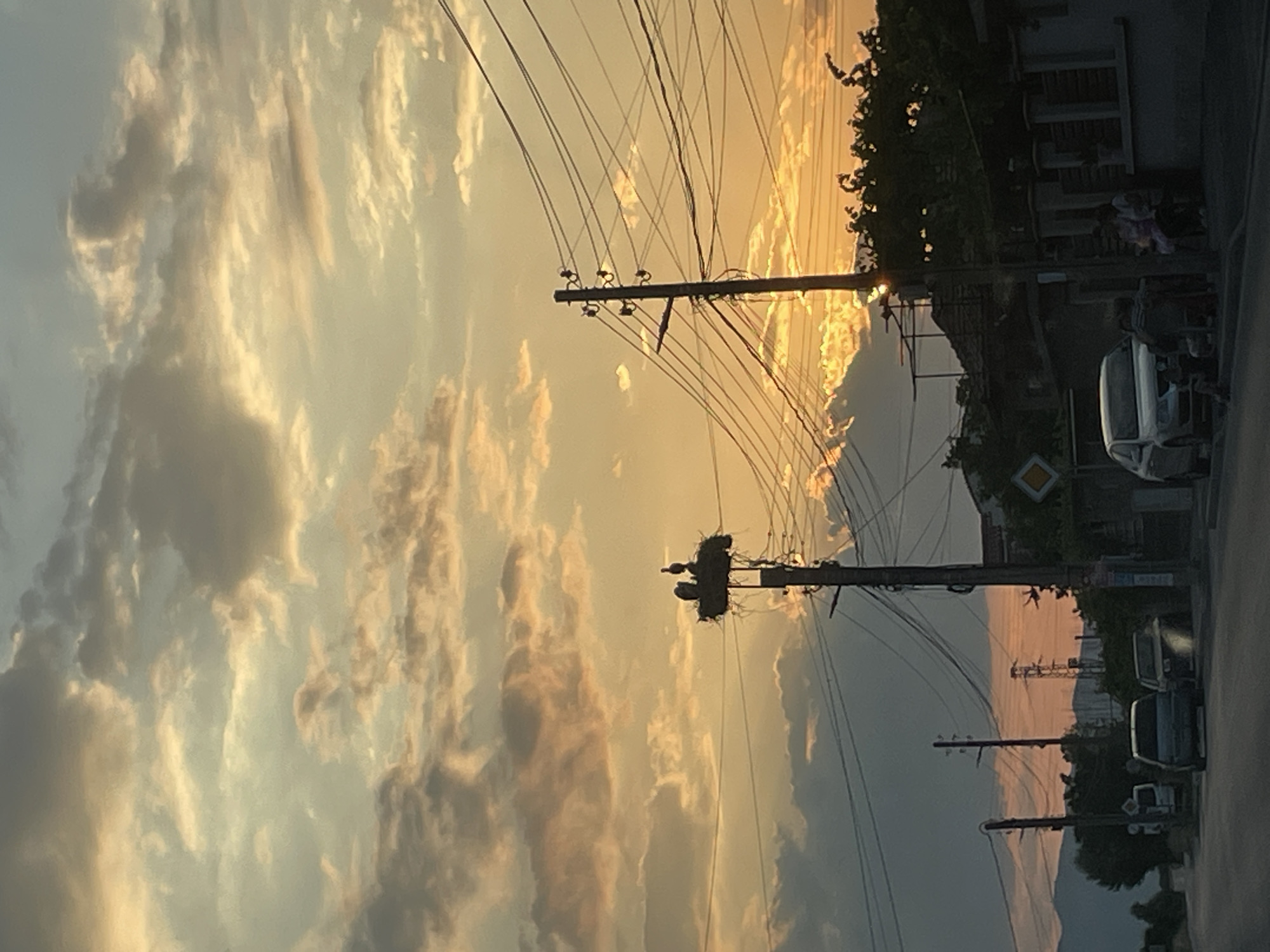 The image captures a sunset scene in an urban or suburban environment. The sky is filled with dramatic clouds tinted with hues of orange, yellow, and gray, indicating the setting sun. In the foreground, a street is visible with several parked cars. Telephone poles with a complex network of wires crisscross the scene, one of which supports a large bird's nest with at least two birds. In the background, there are buildings, trees, and distant mountains or hills. The overall impression is one of a serene, end-of-day atmosphere.