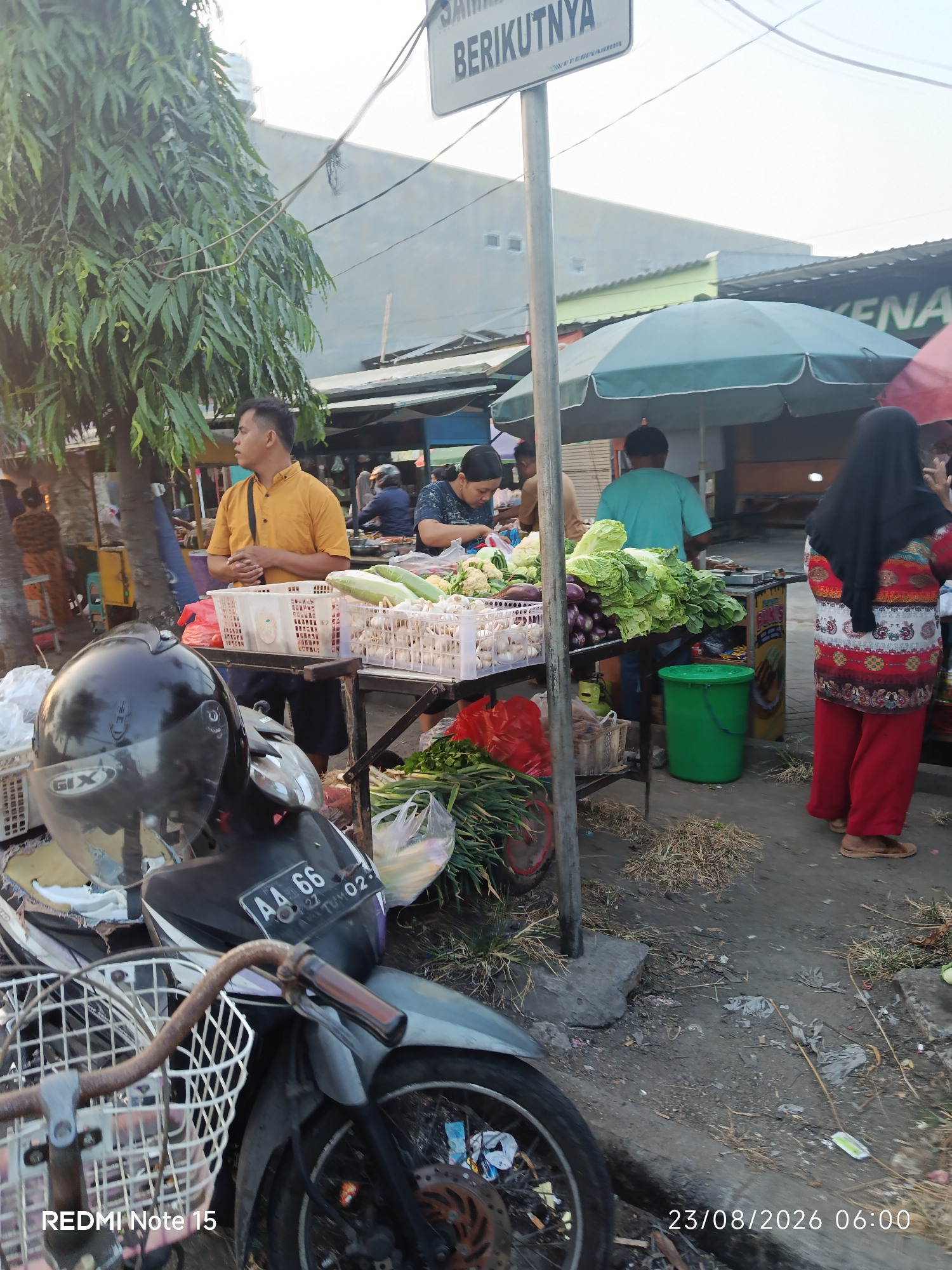 The image captures a street scene, likely a market, in Keboansikep, Indonesia. In the foreground, a motorcycle with a black helmet is parked. To the right, a woman wearing a black hijab and a colorful patterned top with red pants stands near a stall displaying produce. Behind her, various vegetables like cabbage, onions, and gourds are arranged for sale. Several people are visible in the background, some interacting with vendors and others in transit. The environment suggests an outdoor market with stalls and a paved ground surface. A sign with Indonesian text, partially obscured, is mounted on a pole. The timestamp "23/08/2026 06:00" is also present.