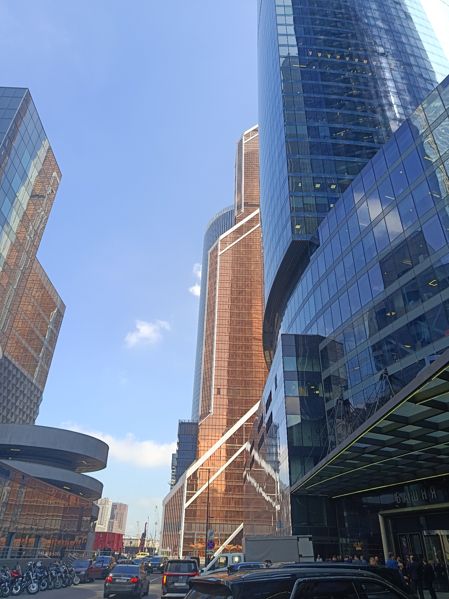 The image shows a street-level view in the Moscow International Business Center (Moscow-City), Russia, characterized by modern high-rise buildings with glass facades. The sky is blue with a few scattered clouds.
Foreground: Cars and motorcycles line the street. People are visible near the entrance of one of the buildings, which features the Russian word "Башня" (tower) above the entrance.
Midground: Several modern skyscrapers are prominently displayed, reflecting the sky and surroundings in their glass surfaces. The buildings are constructed with varying designs and color schemes.
Background: A distant skyline is visible, including what appears to be construction cranes.
Overall: The scene is a typical urban landscape of a modern business district with vehicular traffic and pedestrian activity.