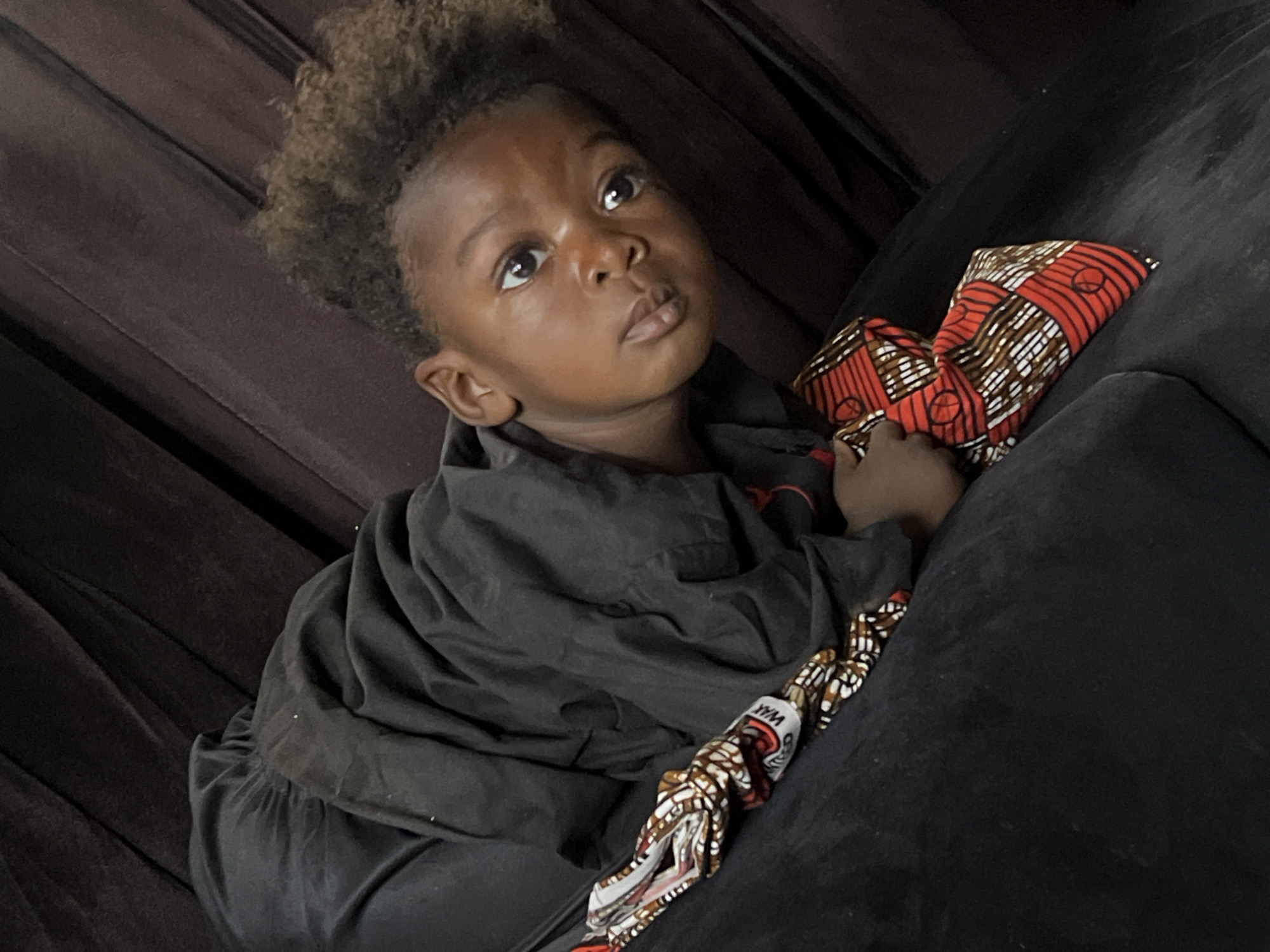 A young child with dark skin and short, curly dark hair is depicted in a close-up, slightly high-angle shot, looking upwards and to the right with a contemplative or curious expression. The child is wearing a loose-fitting, dark (possibly black) long-sleeved garment. They are seated or reclining on a dark, likely upholstered sofa or cushion, which forms the background and foreground, showing subtle folds and textures.
A vibrant, patterned fabric is prominent in the image. The child holds a part of this fabric, and another folded piece rests on the sofa next to them. This fabric displays a striking design primarily in shades of red and orange, accented with white and brown geometric and circular patterns. Upon close inspection, the word "WAX" is faintly visible on a segment of the fabric, suggesting it is a type of African wax print.
The scene appears to be indoors, characterized by soft, somewhat dim lighting that creates subtle highlights on the child's face and eyes. The setting, according to context, is Jos, Nigeria. The child's gaze conveys a sense of quiet observation or perhaps a moment of wonder.