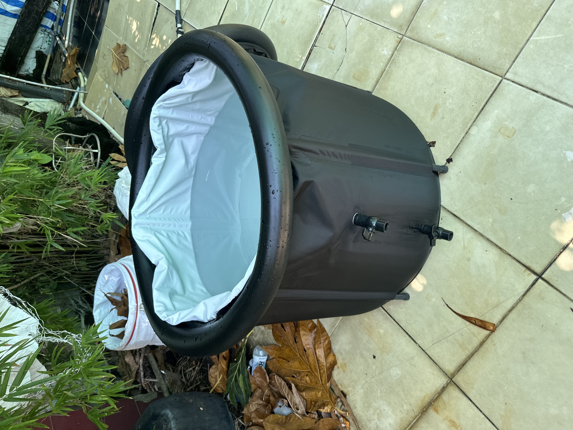 A black, collapsible, inflatable bathtub filled with clear water sits on a tiled patio in Nha Trang, Vietnam. The bathtub has a white liner and a spigot at the bottom. Surrounding the bathtub are fallen leaves, some bamboo plants, and other debris. The scene appears to be outdoors, possibly in a backyard or courtyard.