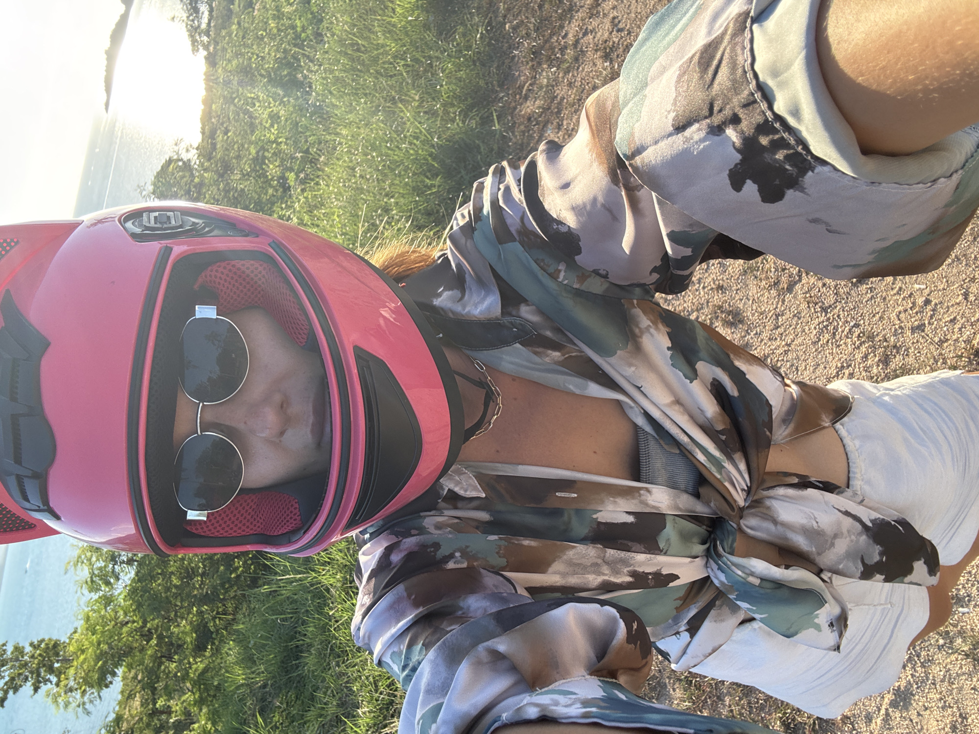 A woman is taking a selfie outdoors, wearing a pink helmet with cat ears, sunglasses, and a patterned shirt tied at the waist over white shorts. The background shows a body of water, greenery, and a bright sky with the sun casting a glare. The terrain appears to be dirt and grass. The location is Unknown, Unknown.