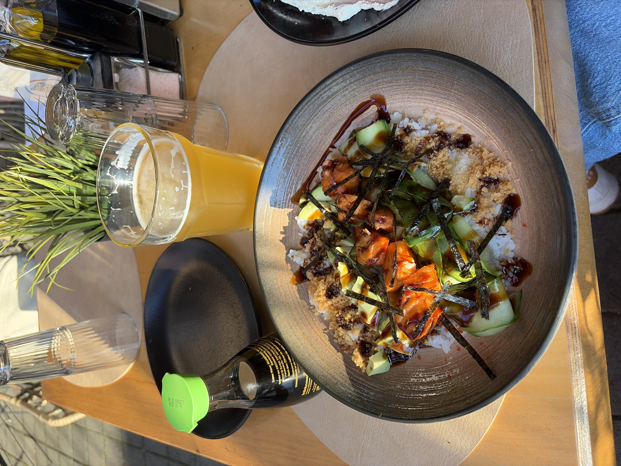 This image captures a close-up of a meal served on a wooden table, likely in an outdoor or brightly lit indoor setting, in Unknown, Unknown. The central focus is a stoneware bowl with a dark, textured rim, containing a dish composed of white rice topped with diced seared salmon, sliced avocado, sliced cucumber, strips of dark green nori seaweed, a drizzle of dark reddish-brown sauce, and light brown crunchy particles.
To the left of the bowl, a small black side plate is placed next to a clear glass bottle of Kikkoman soy sauce, which has a bright green cap displaying "15.01.2023 L256 10:40" and German text on its label. To the right, a faceted clear glass holds a light yellow, foamy beverage, resembling beer. An inverted, ribbed clear glass stands beside it. In the background, a metal caddy holds multiple condiment bottles, including clear bottles likely for oil and vinegar, and two shakers, one containing pink salt. A green artificial plant in a dark pot is positioned behind the glasses.
The wooden table is partially covered by a light tan placemat. Strong, natural sunlight illuminates the scene from the left, creating distinct shadows. Part of a wicker chair is visible in the upper left background, and a person's leg, clad in blue jeans and a white shoe, is partially visible under the table in the lower right.