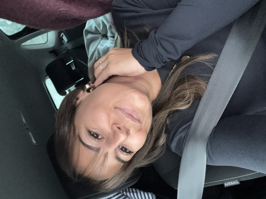 A female individual is seated in the front passenger seat of a vehicle in Valence, France. She has medium-length brown hair with lighter highlights and bangs, and is looking directly at the viewer with a slight smile. She wears a dark, long-sleeved top, a gold-colored circular pendant necklace, and prominent gold-colored hoop earrings. A silver ring is visible on the ring finger of her left hand, which is raised and positioned near her right ear. A grey seatbelt is securely fastened across her chest and lap, displaying an "AIRBAG" label on the seat fabric to her left. The vehicle's interior includes a black fabric seat and a black headrest. In the background, beyond the seat, blurred forms suggest a light grey blanket or garment and a dark red or maroon textured object. The exterior visible through the vehicle's window appears overcast and indistinct.